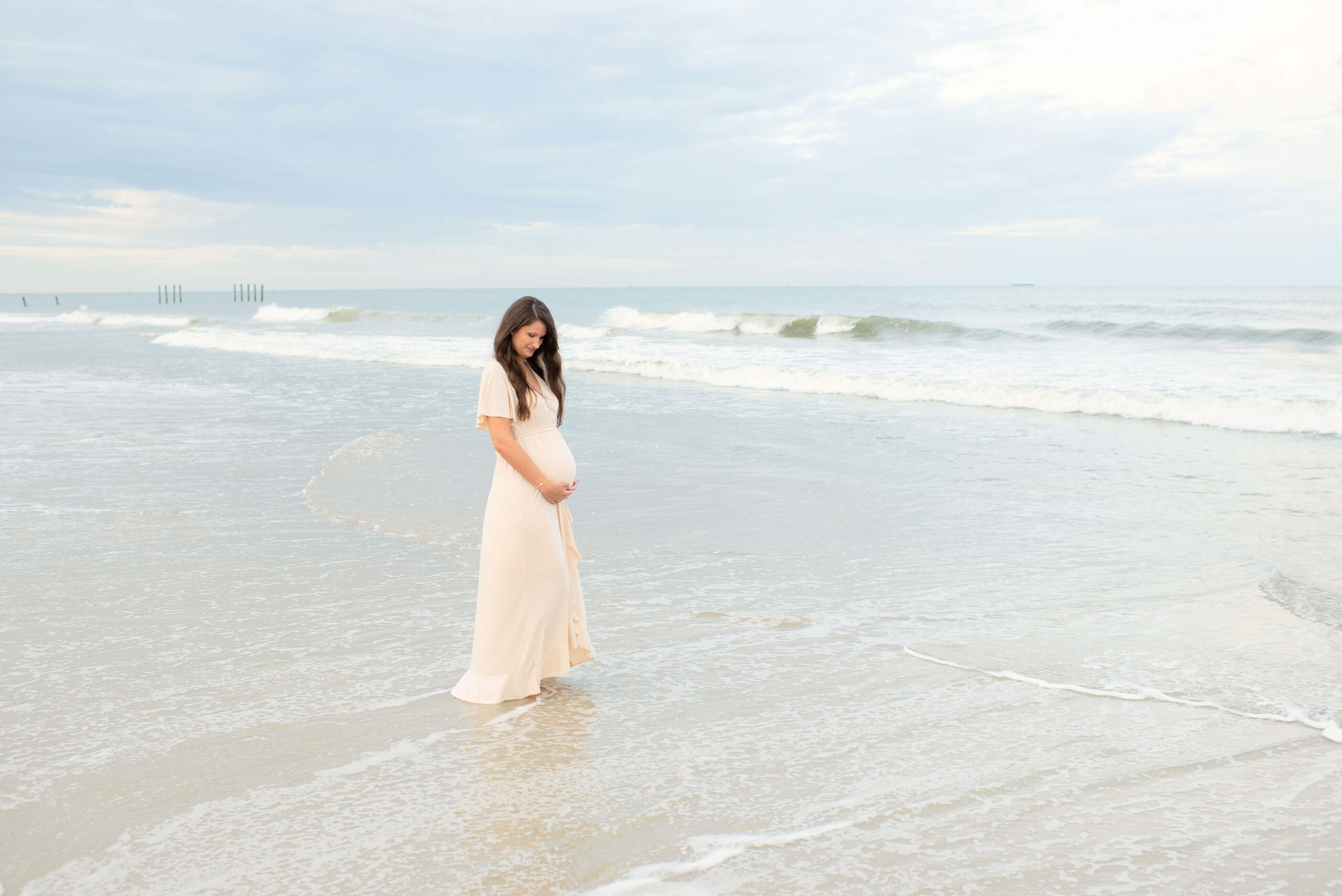A mom to be stands on the beach in a beige maternity dress while holding her bump Jacksonville midwives
