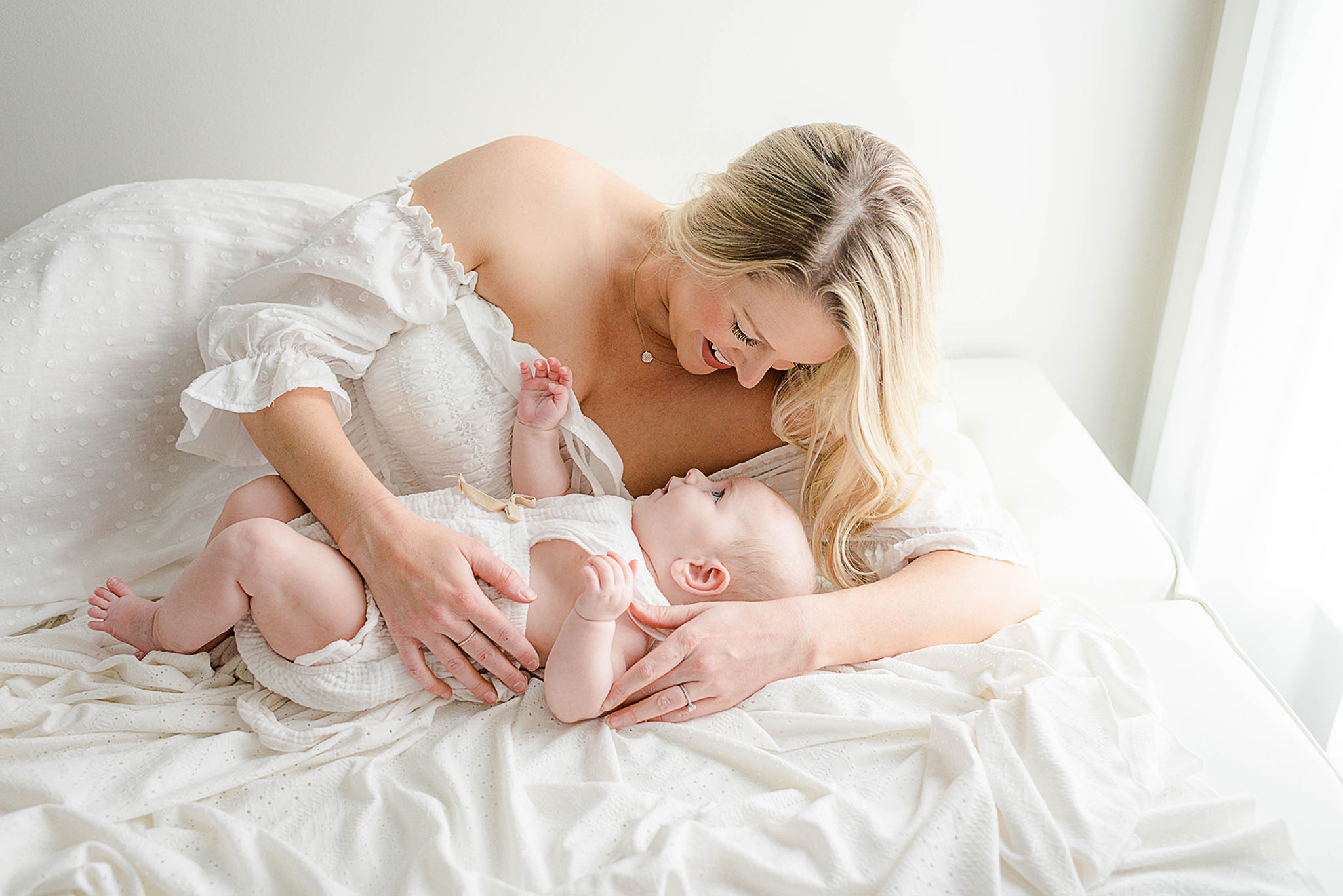A mom in a white dress lays on a bed cuddling her infant baby in a white onesie