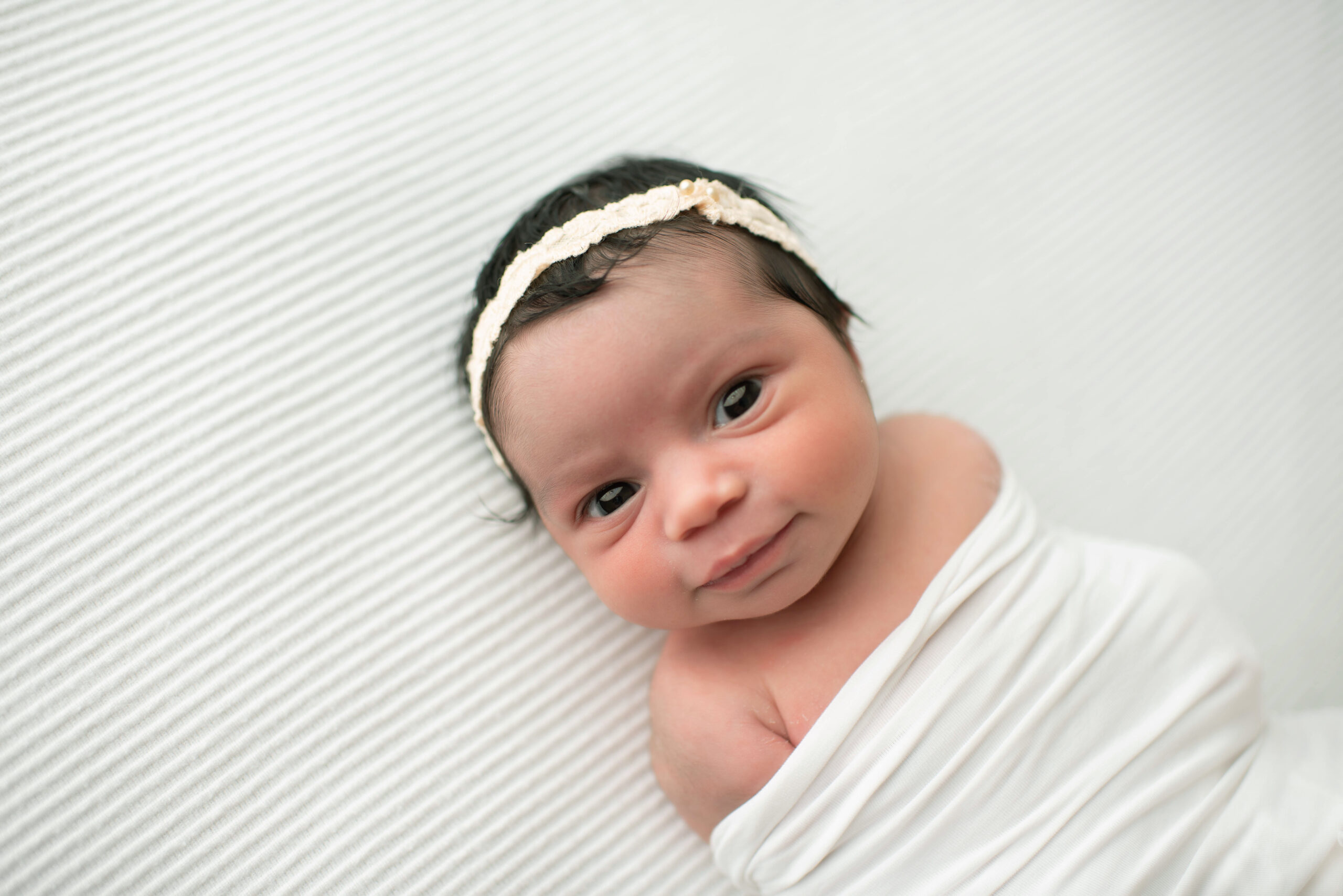 A newborn baby lays on a white bed in a headband