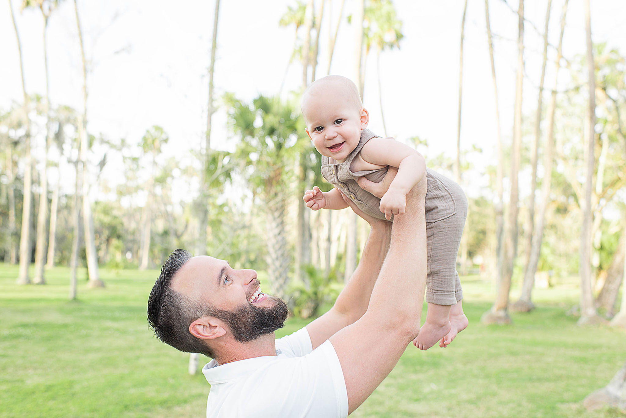 A father lifts his infant son wearing brown overalls over his head while playing in a grassy field jacksonville nannies