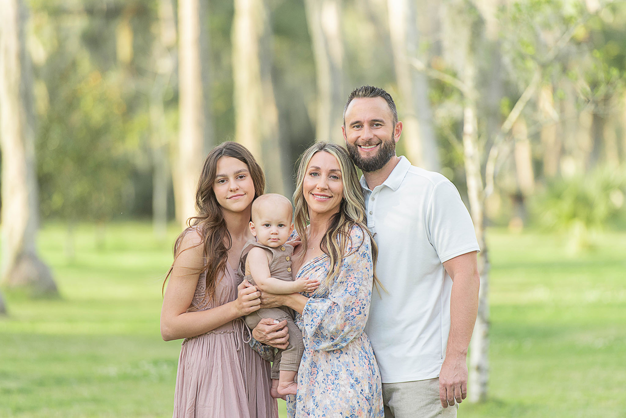 A mother stands in a grassy field of palm trees holding her infant son while her husband and teenage daughter hug onto them