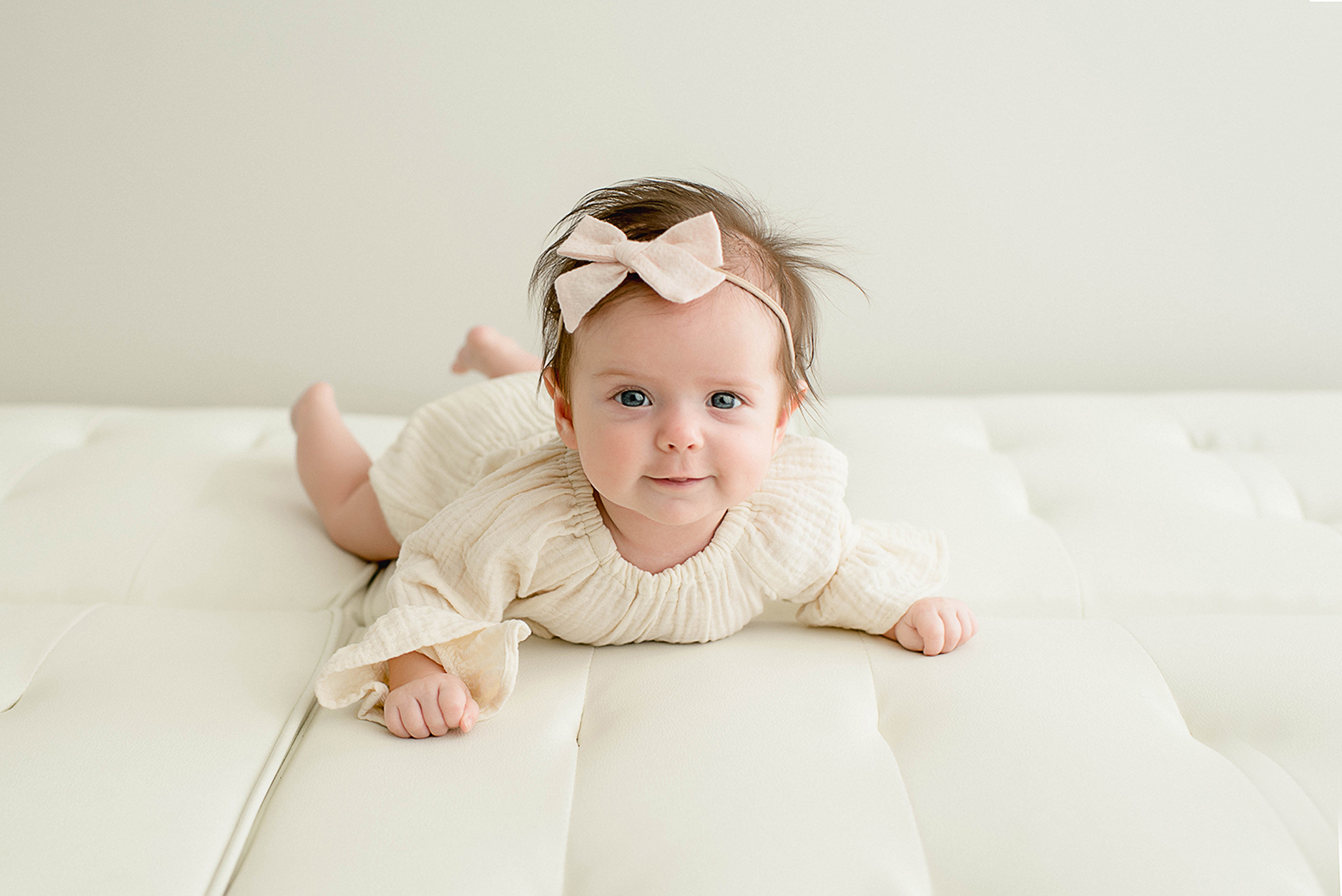 An infant girl has belly time on a leather couch in a white dress and bow lactation consultant jacksonville fl