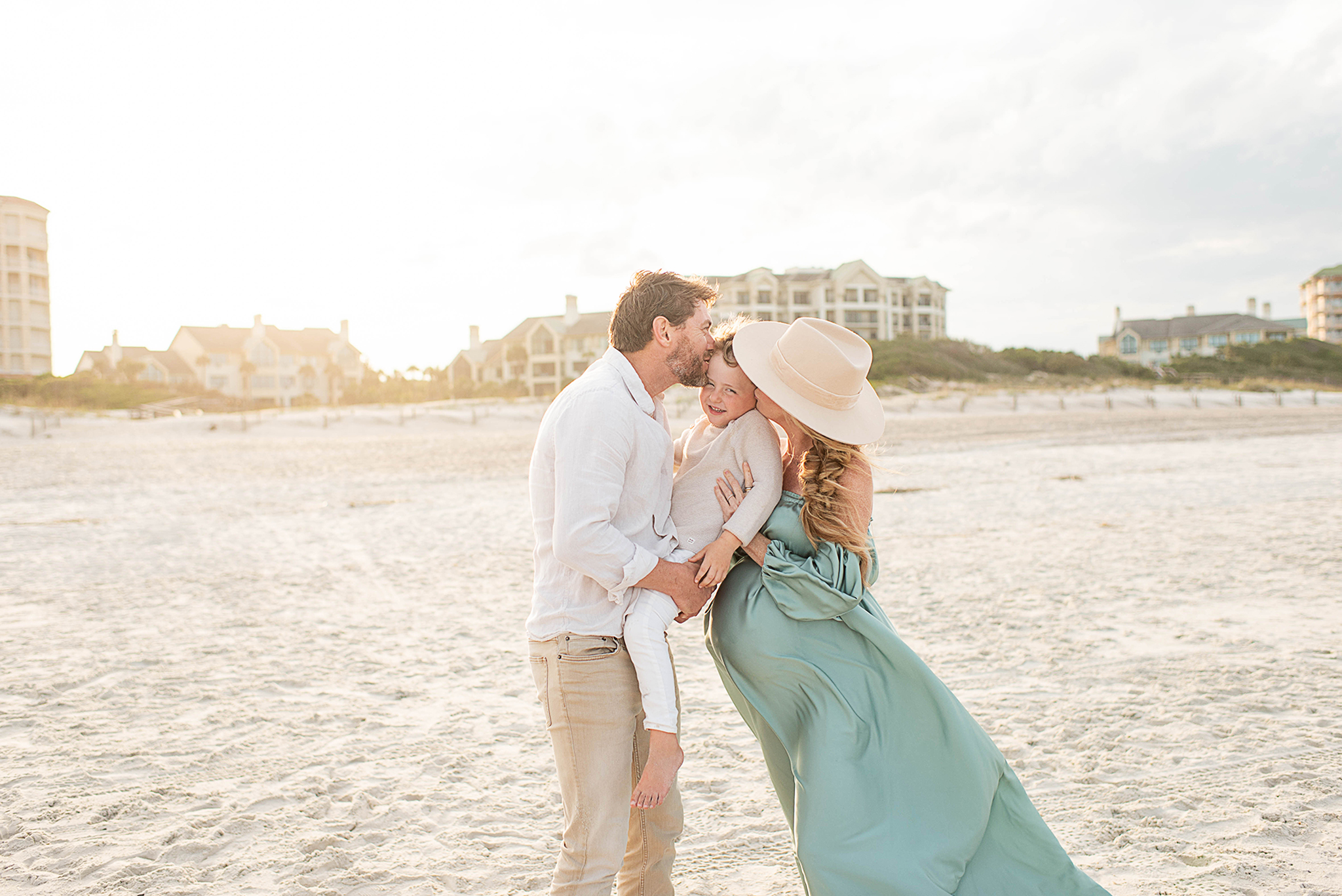 pregnant woman in green flowy dress kissing son with man kissing son on the jacksonville beach