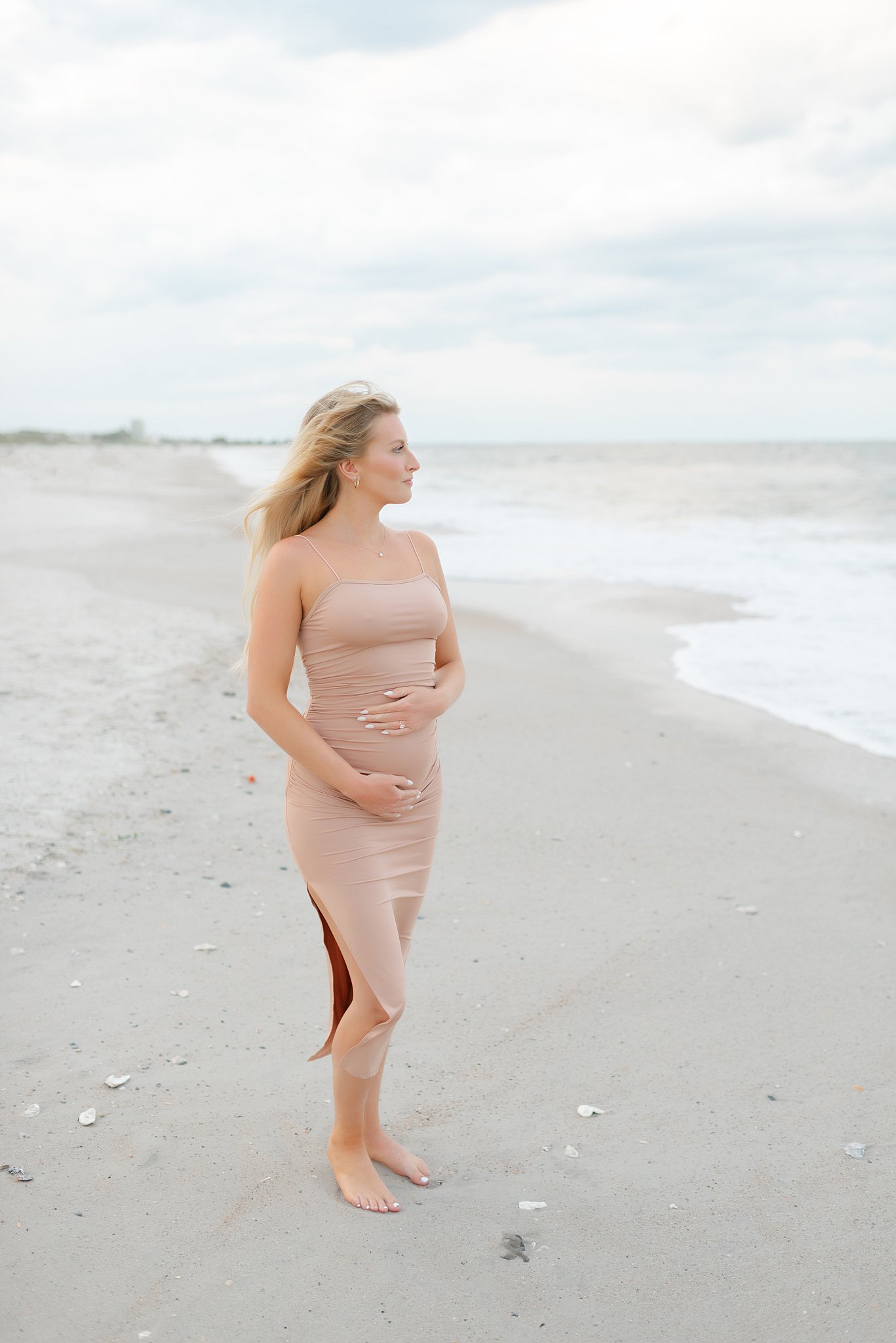 A mother to be gazes to the ocean while standing in a beige maternity dress after her 3d ultrasound Jacksonville