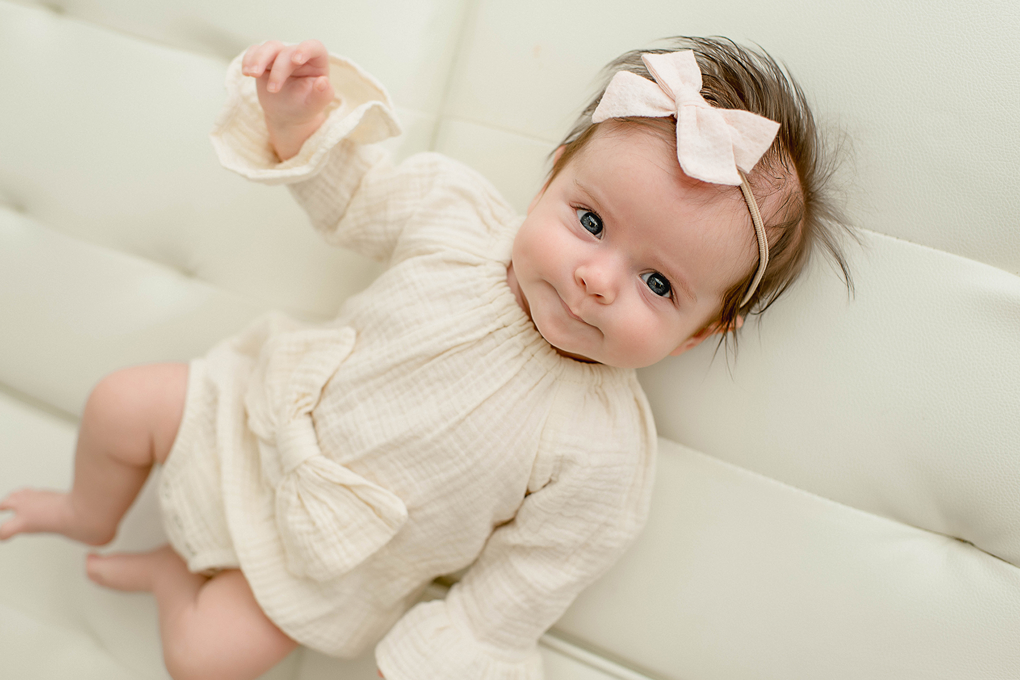 A smiling toddler girl in a cream onesie and matching bow lays on a leather bench with an arm up