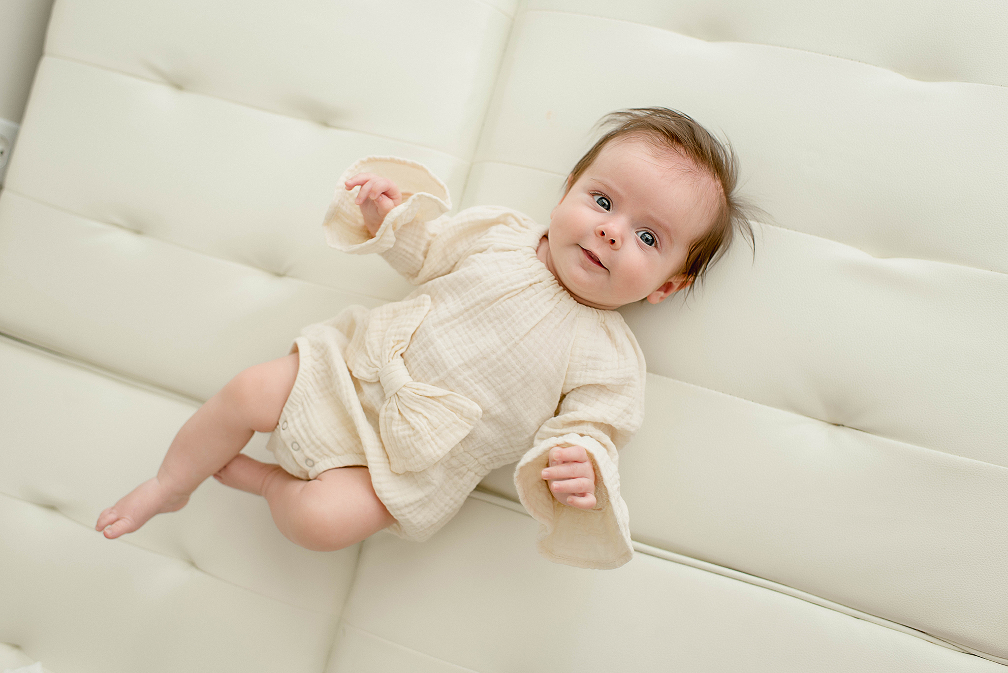 An infant baby lays on a leather couch smiling in a cream onesie before visiting daycares in jacksonville