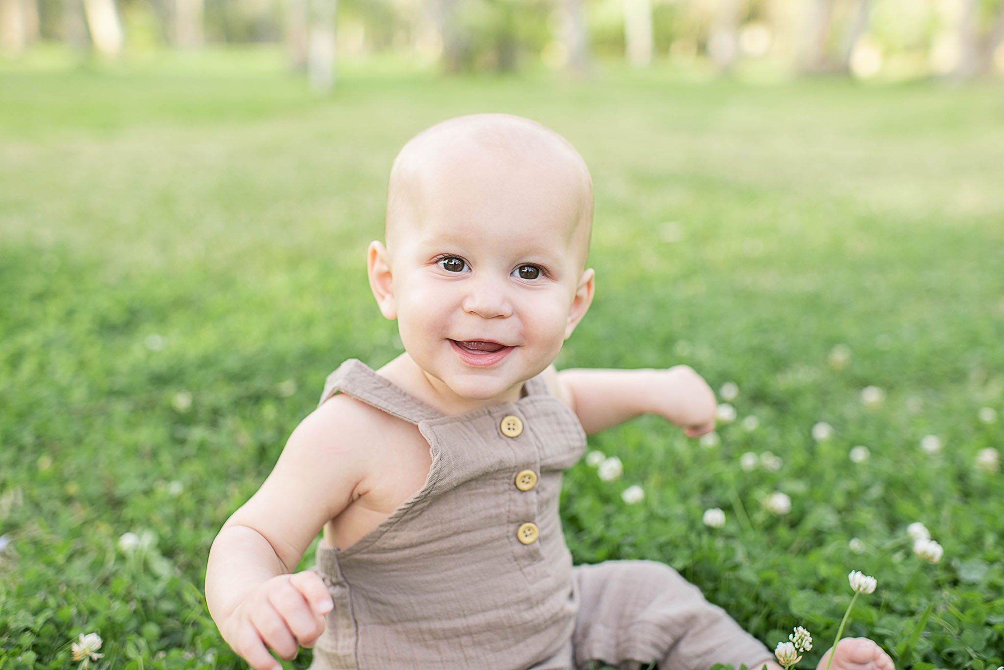 A happy toddler boy in a brown onesie sits in a field of clover smiling after visiting a jacksonville pediatric dentist