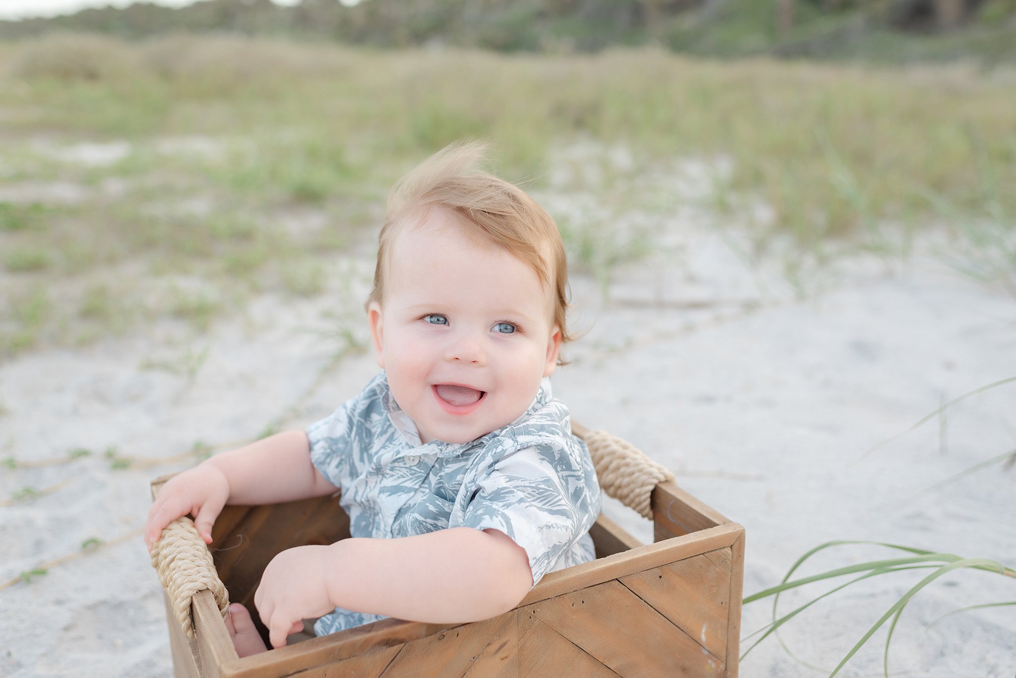 A smiling toddler boy sits in a wooden box on a windy beach
