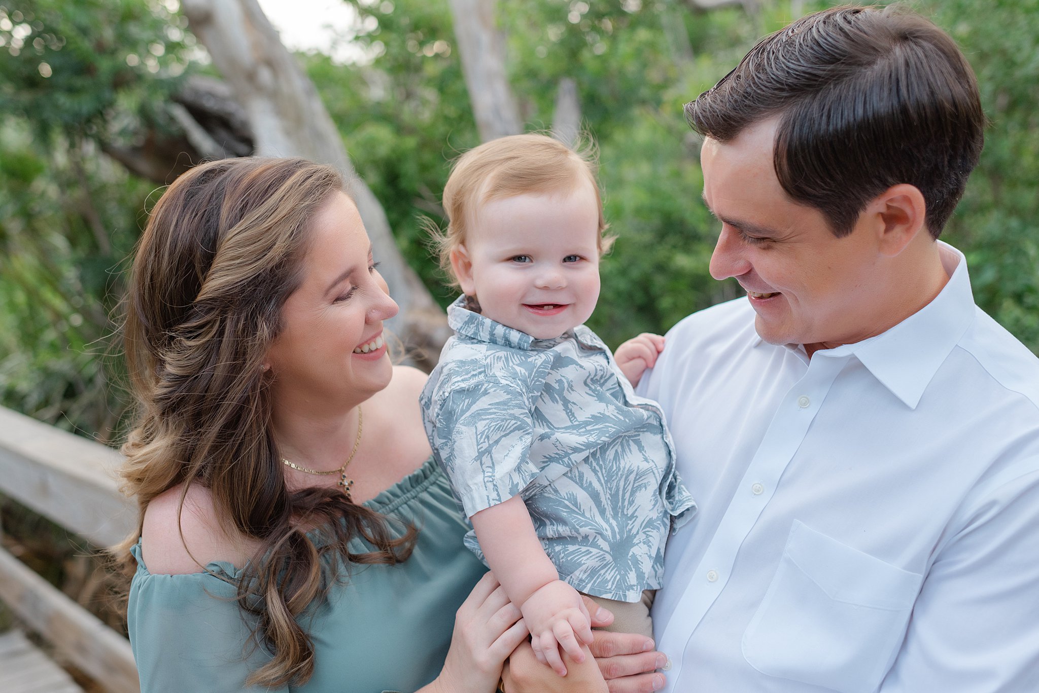 A happy toddler boy sits in dad's arms on a beach boardwalk with mom smiling on after visiting jacksonville pediatricians