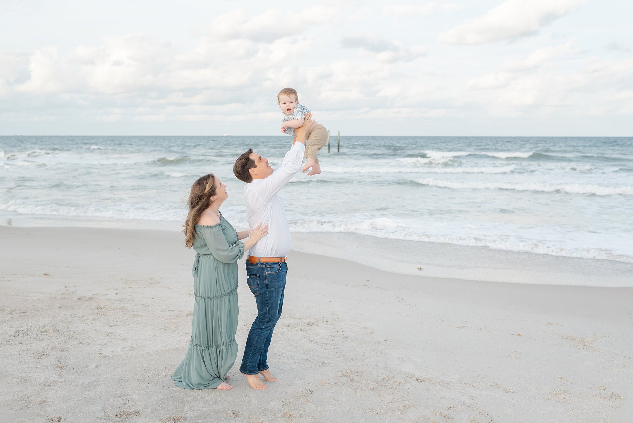 A father lifts his happy infant above his head while standing on a windy beach with mom at his side after visiting jacksonville pediatricians