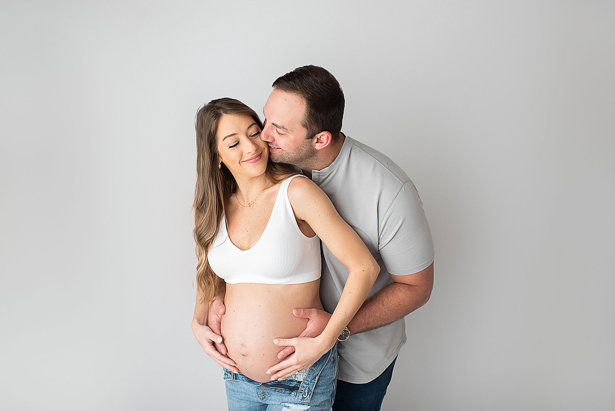 A husband holds his pregnant wife's bump while standing in a studio in jeans before she gets a jacksonville prenatal massage
