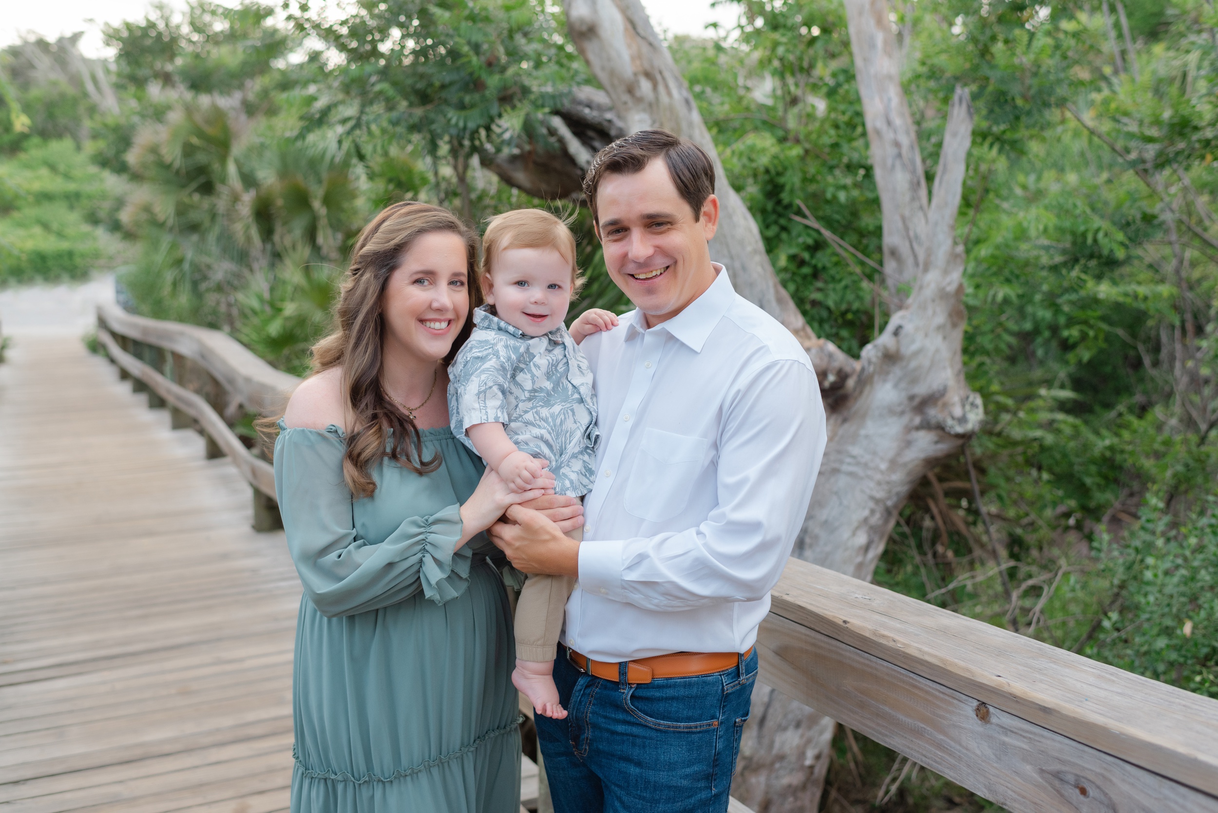 A happy toddler sits in dad's arms with mom and dad standing on a boardwalk after visiting Children's Clothing Stores Jacksonville FL