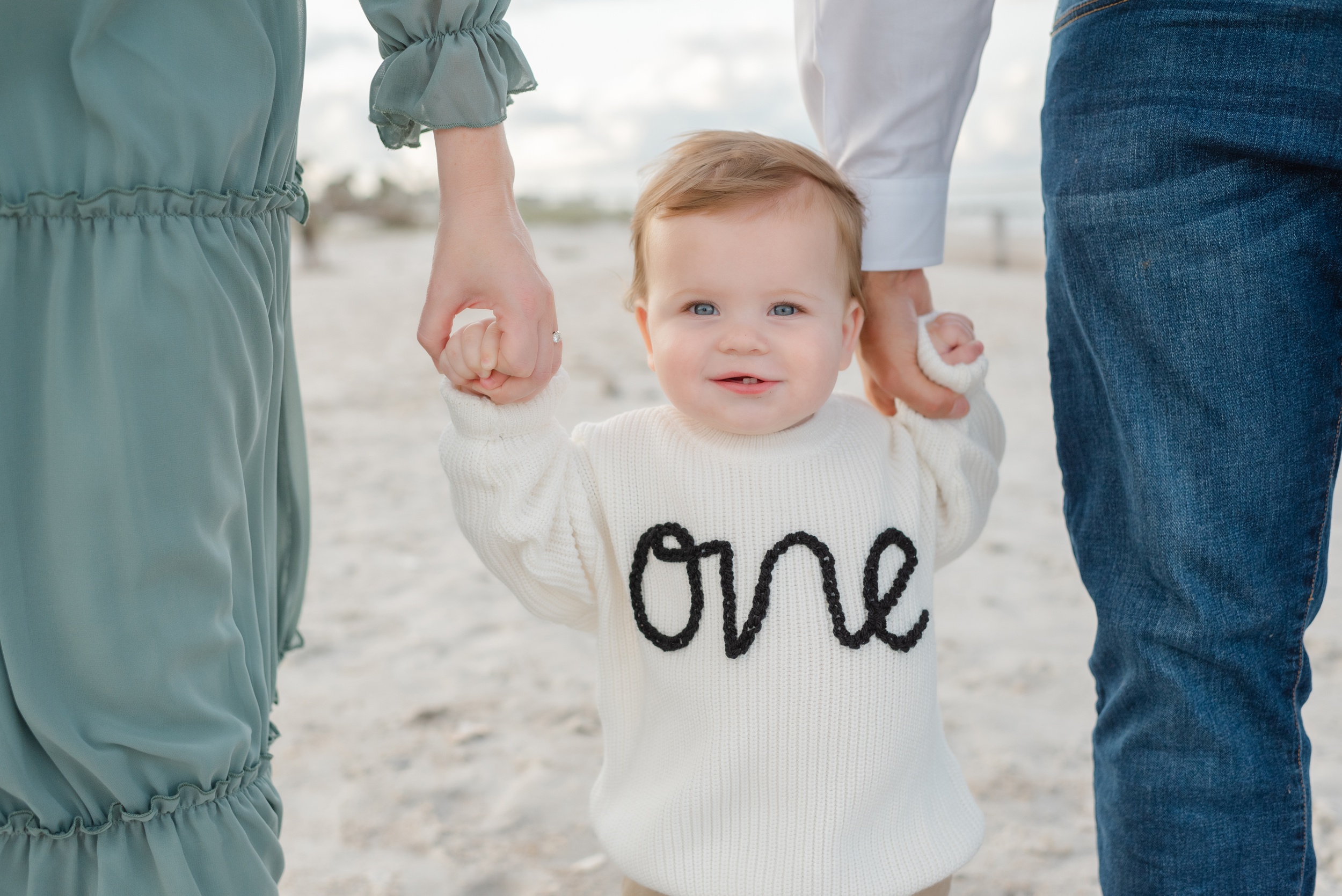 A young toddler walks on a beach holding mom and dad's hands in a white sweater from Children's Clothing Stores Jacksonville FL