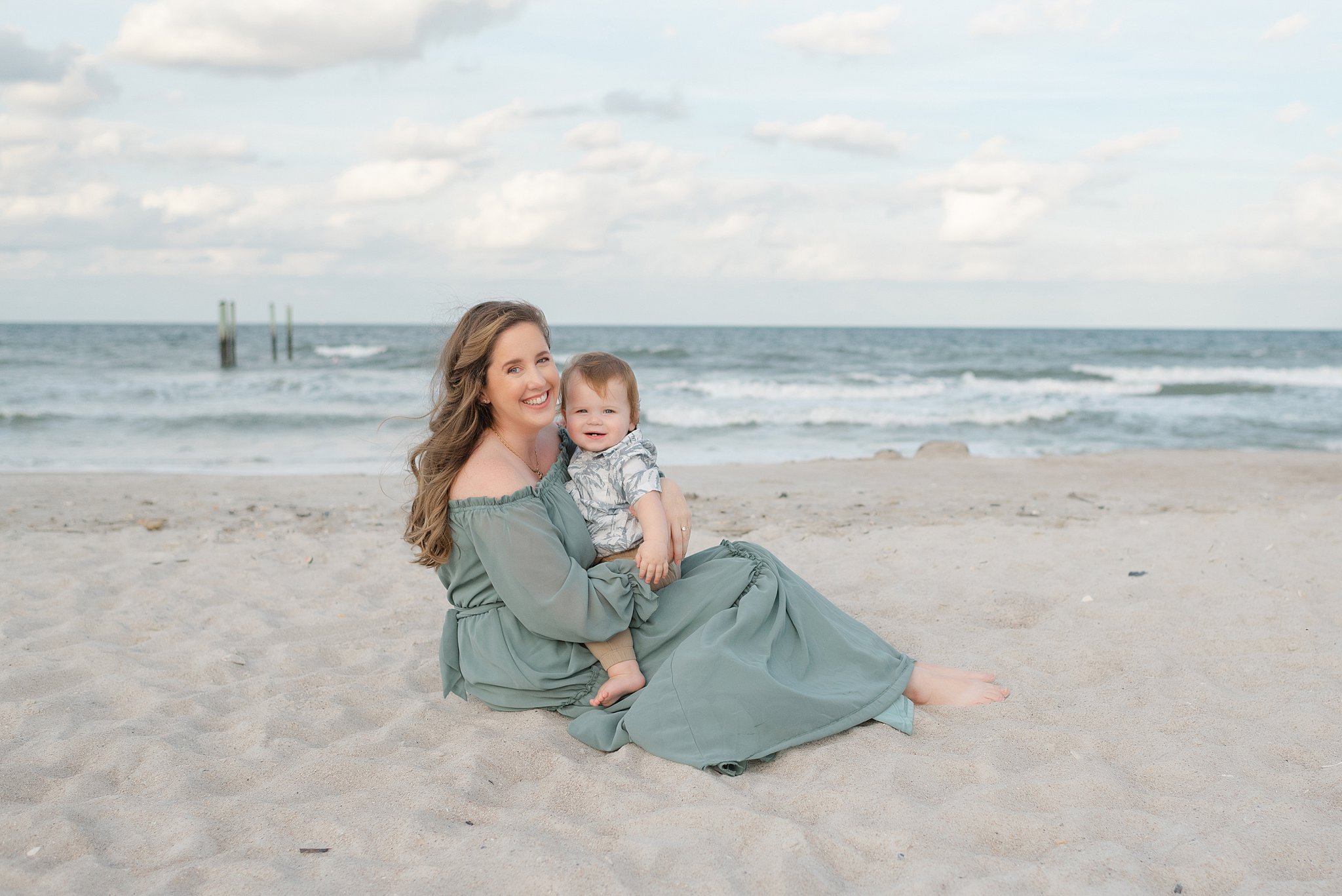 A happy mother in a green dress sits on a beach with her toddler son in her lap