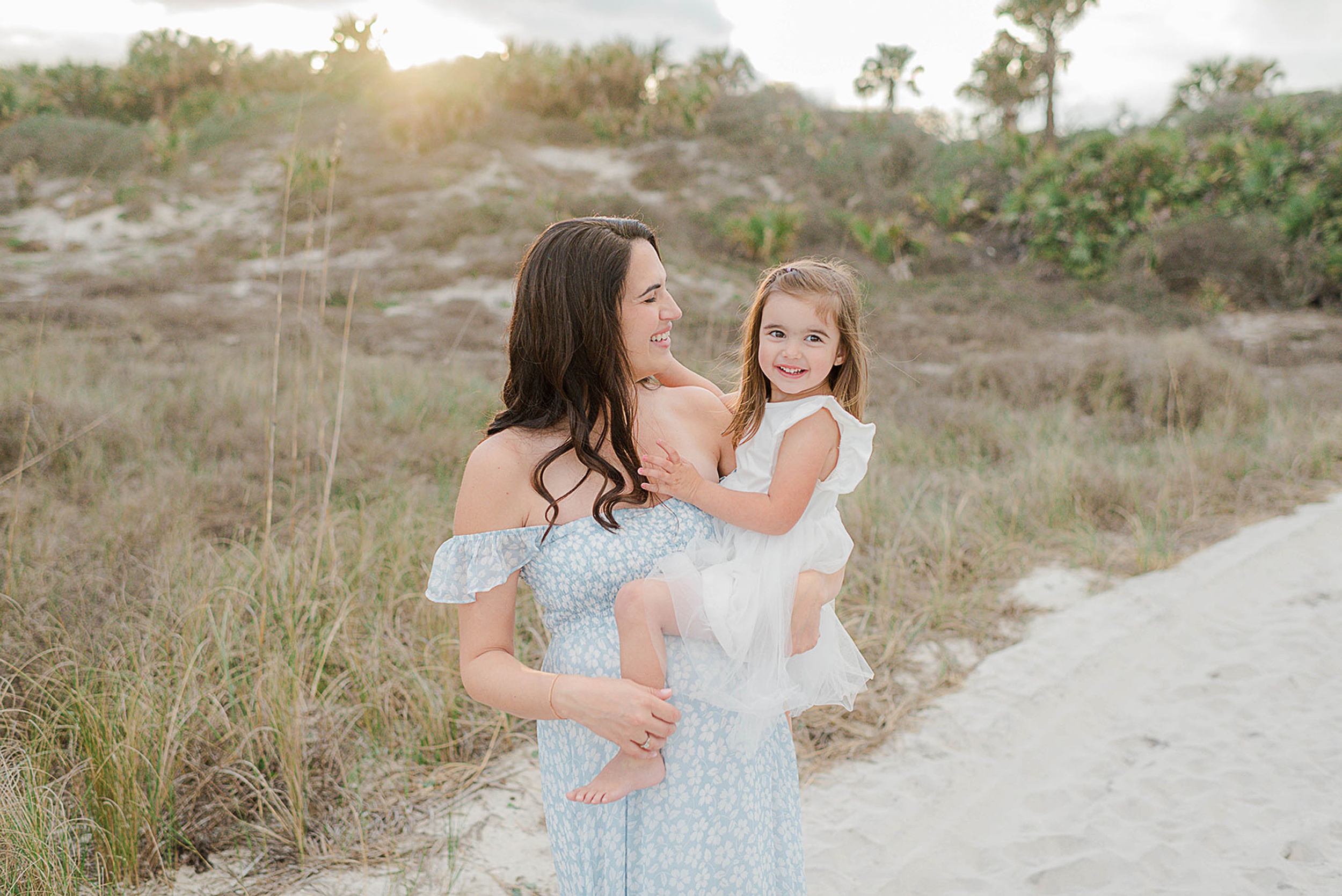 A mother in a blue dress walks on a beach path with her toddler daughter on her hip in a white dress