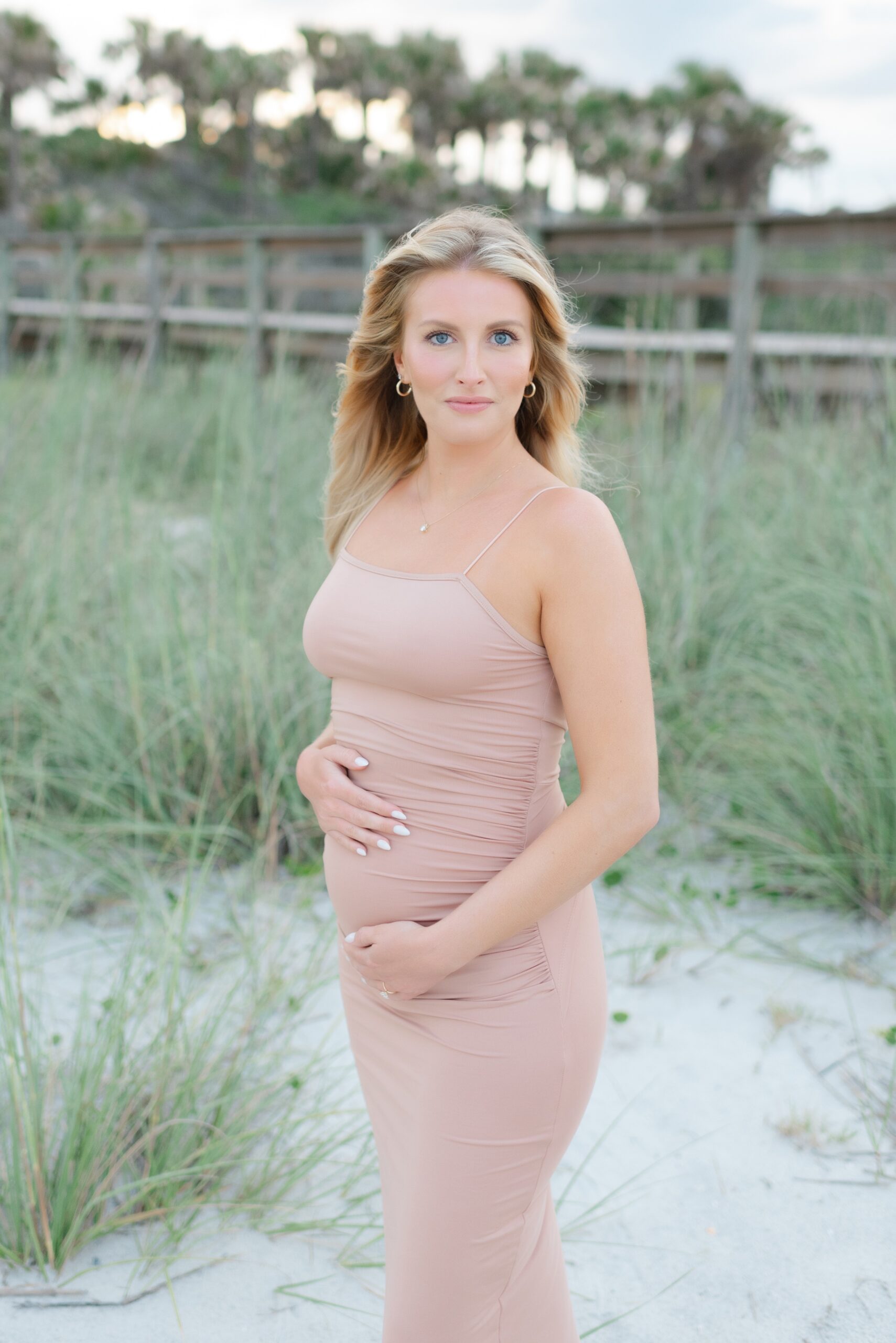 A mother to be stands in a beach dune holding her bump in a pink dress before exploring pelvic floor therapy jacksonville fl