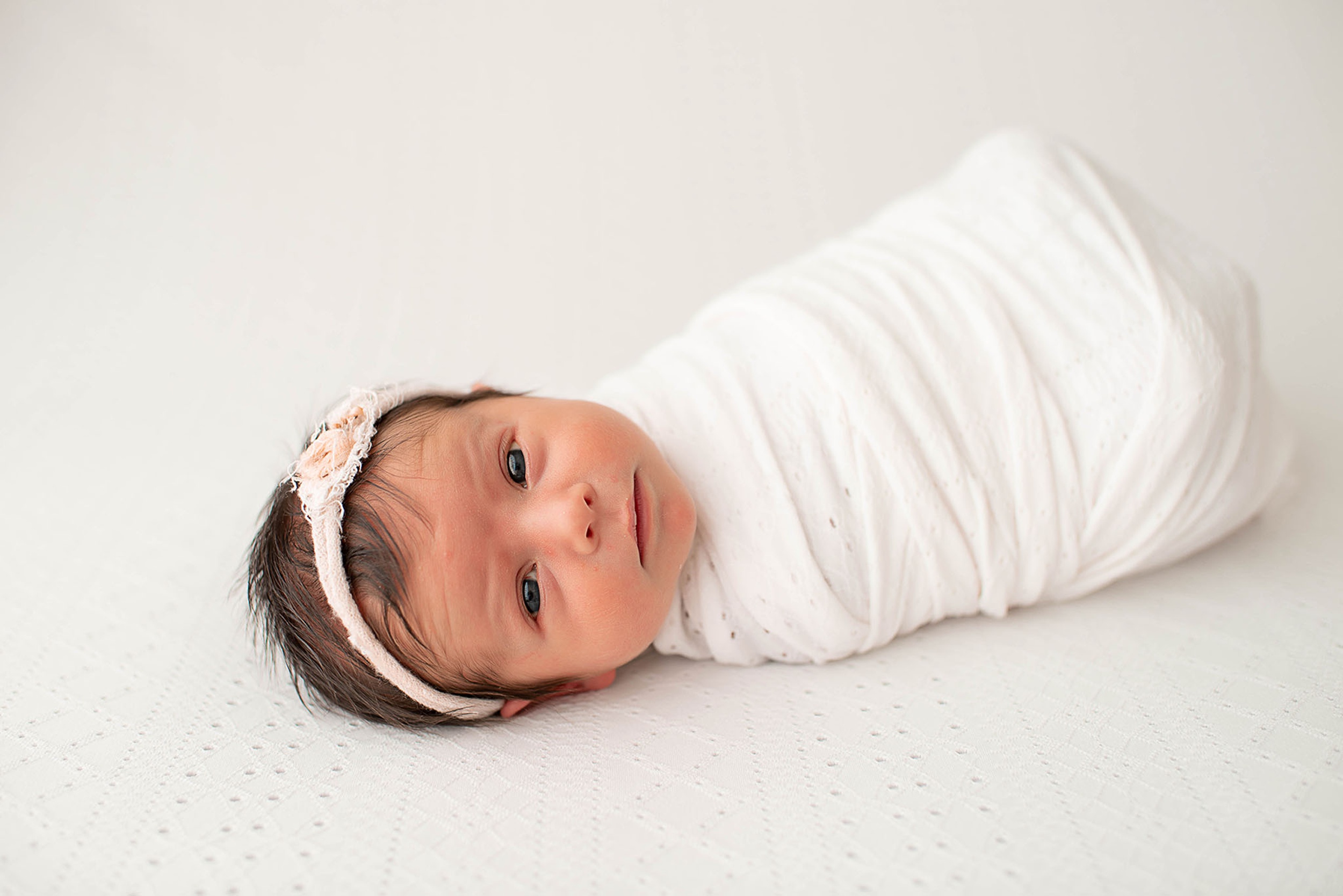 A newborn baby girl lays in a white swaddle on a matching blanket with eyes open