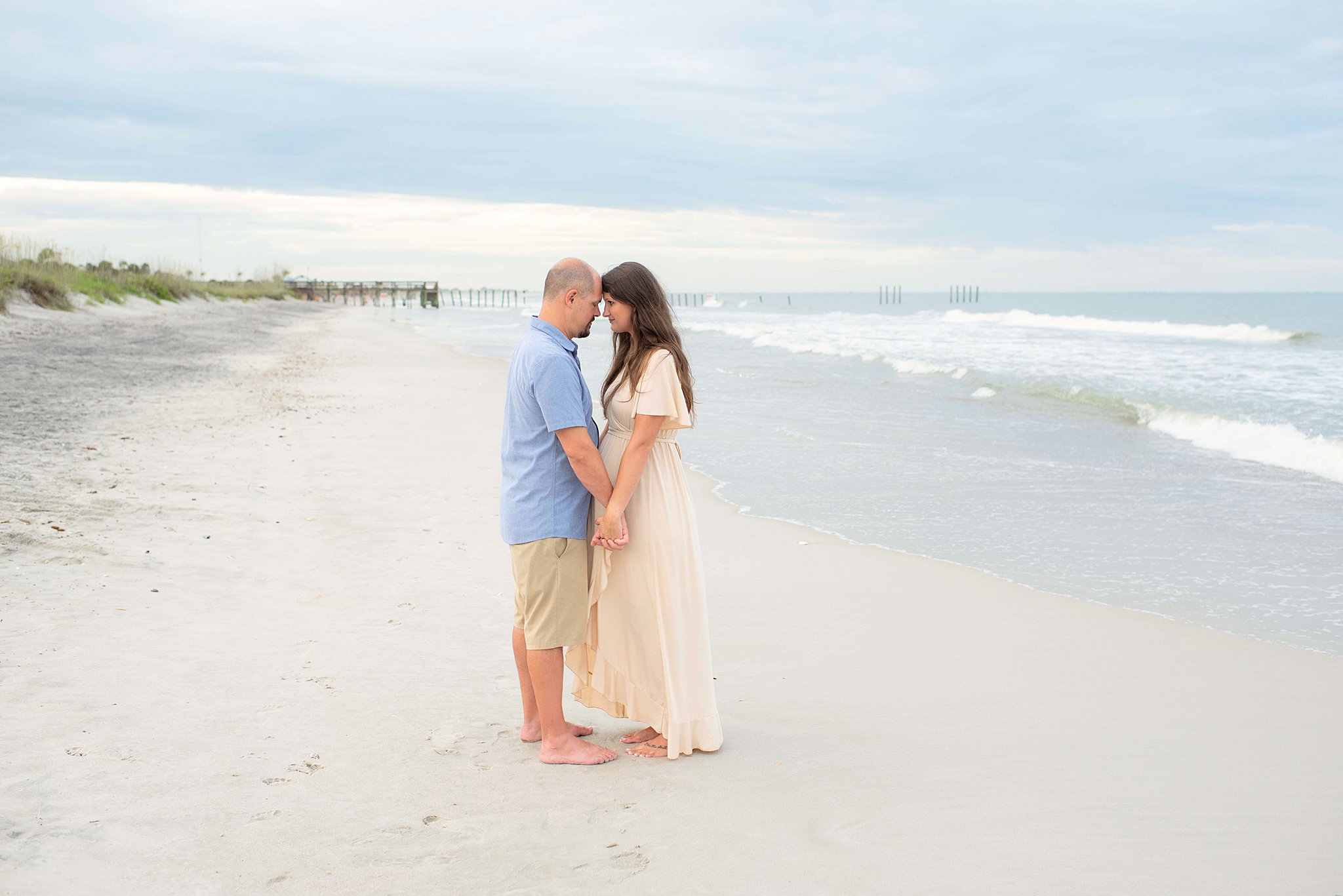 Expecting parents stand on a beach holding hands with foreheads touching