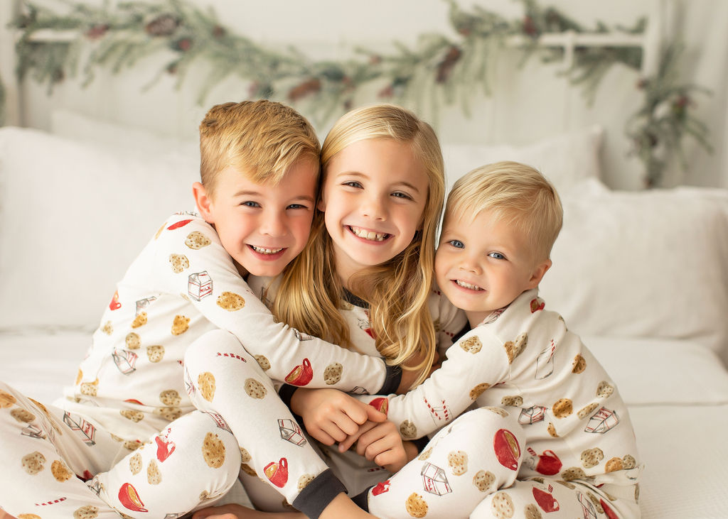 Three toddler siblings smile and hug each other in matching pajamas while sitting on a bed in a studio during some christmas mini sessions jacksonville fl