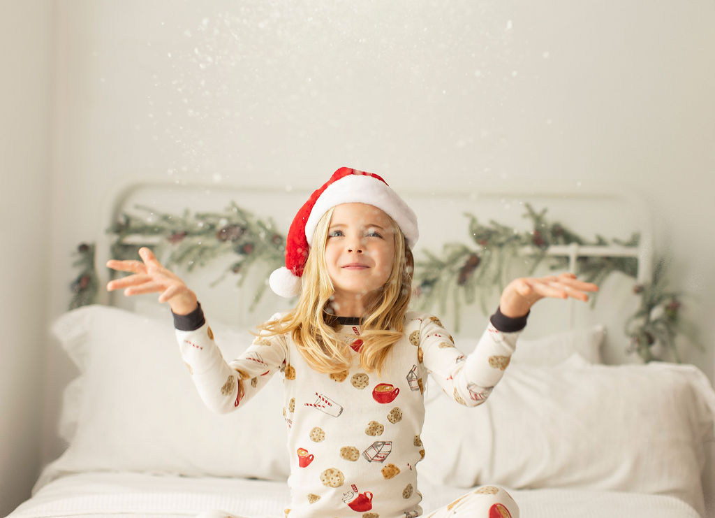 A young girl tosses glitter over herself while sitting on a bed in pajamas and a santa hat