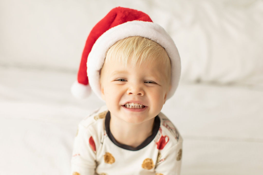 A toddler boy sits on a bed in pajamas smiling big with a santa hat on during christmas mini sessions jacksonville fl