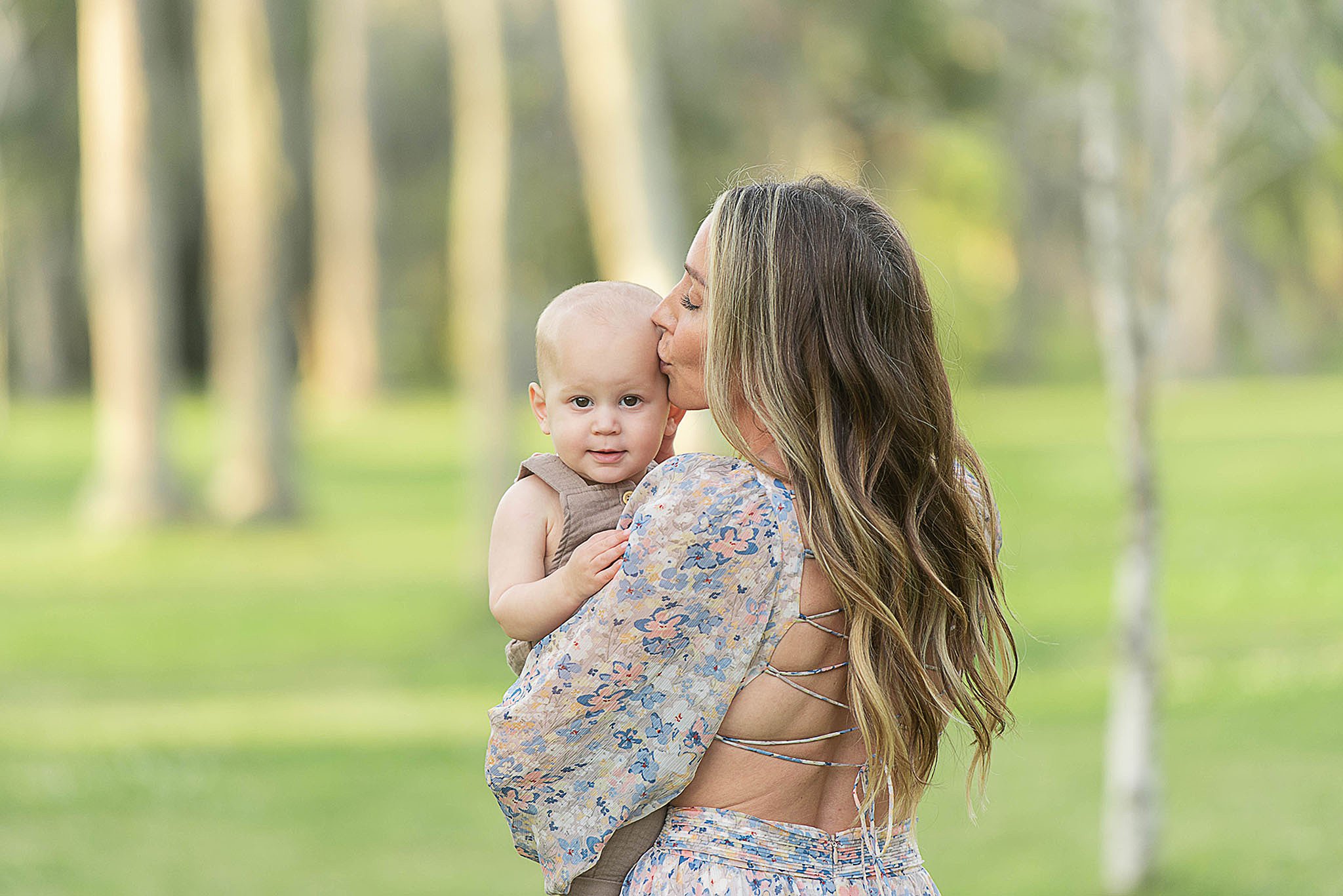 A mother in an open back floral print dress kisses the head of her infant in her arms while standing in a park at sunset Mommy And Me Classes Jacksonville FL