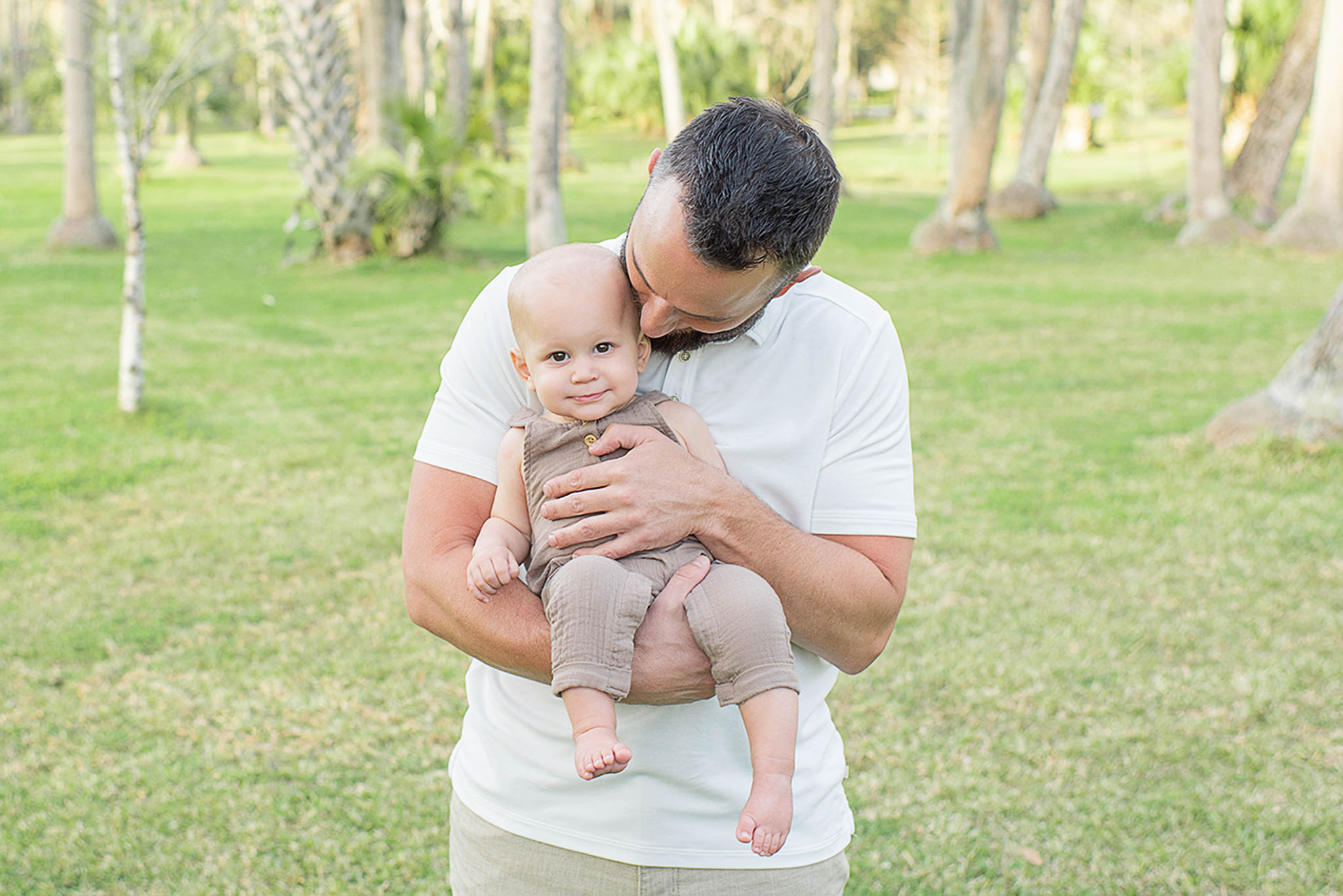 A happy father stands in a park kissing the head of his toddler son in brown overalls