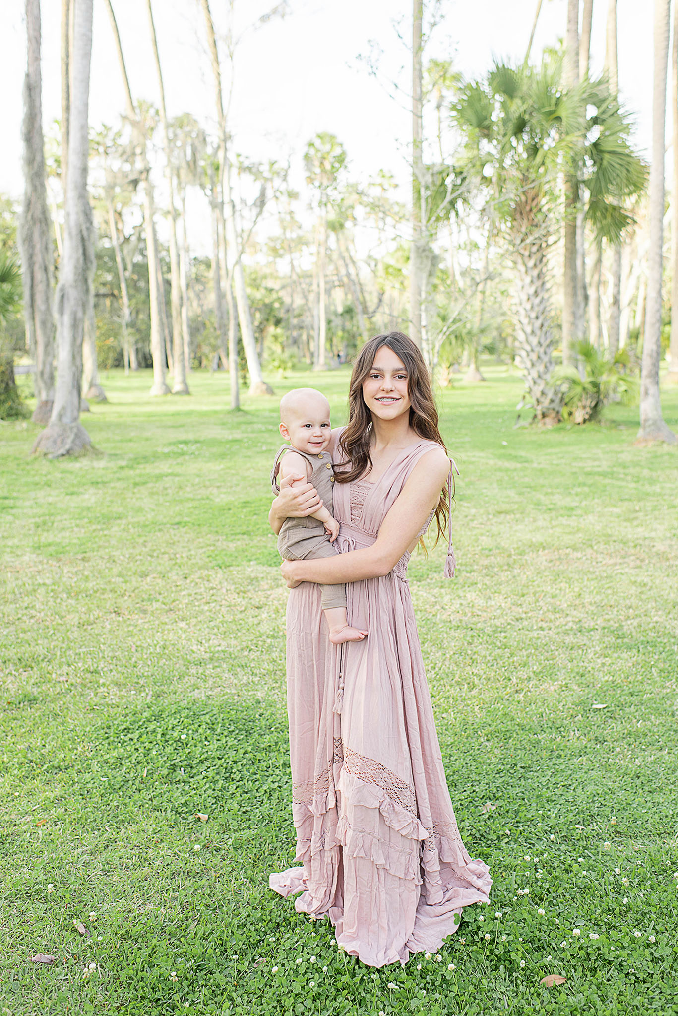 A proud teenage sister stands in a park holding her infant baby brother on her hip in a pink dress during things to do with toddlers in jacksonville fl