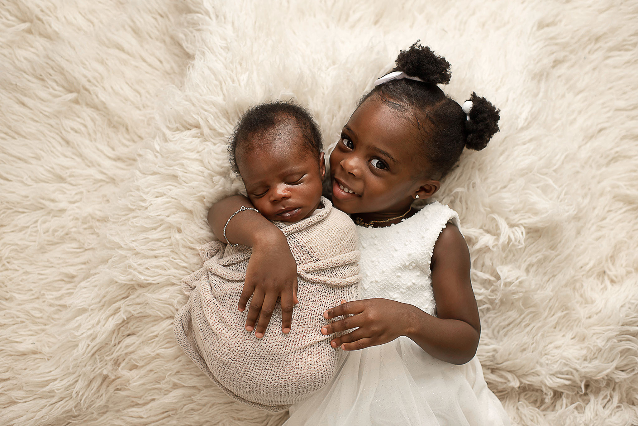 A toddler girl ina. white dress lays on a fur blanket while snuggling with her newborn baby sibling