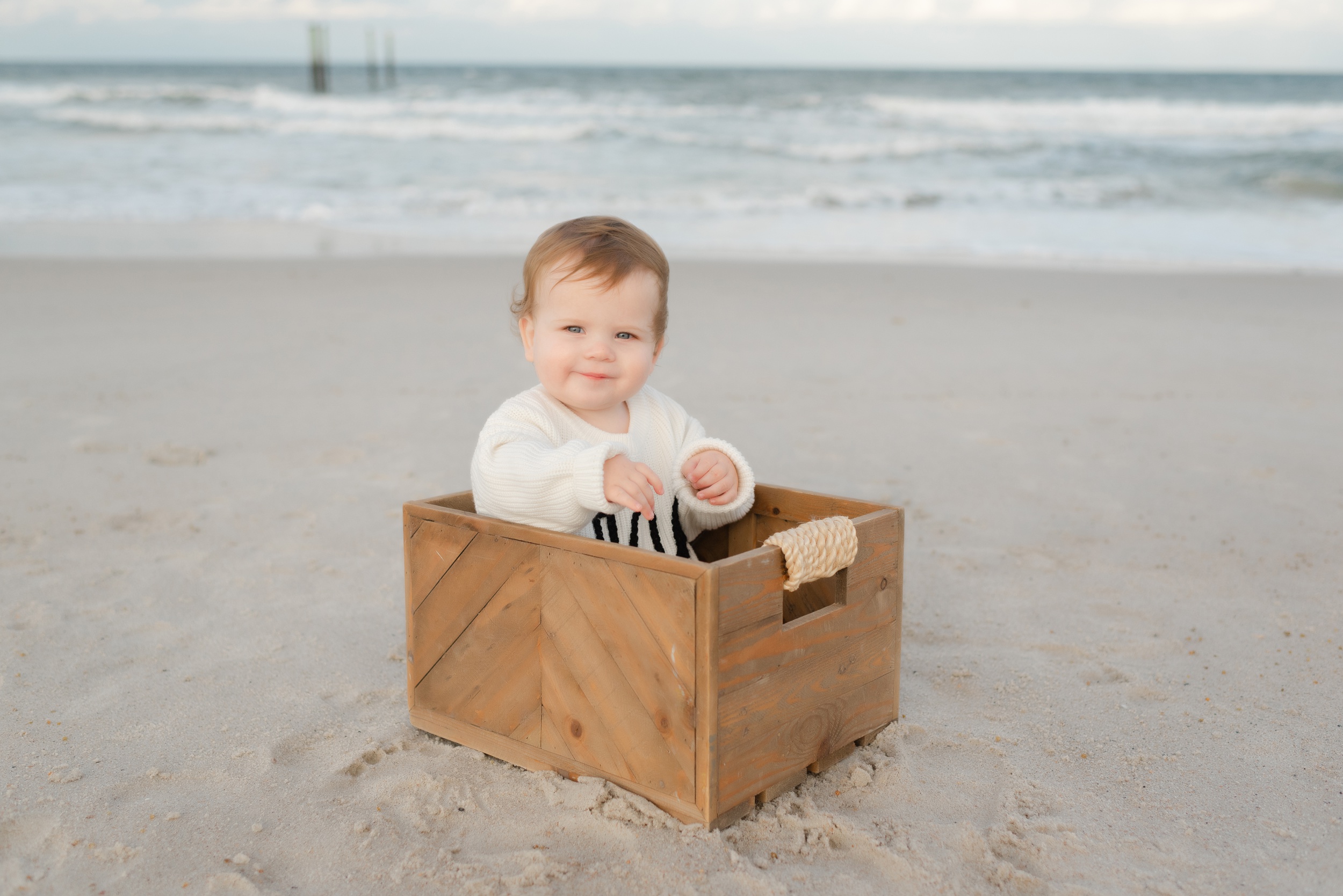 A toddler boy sits in a wooden box on the beach in a white sweater