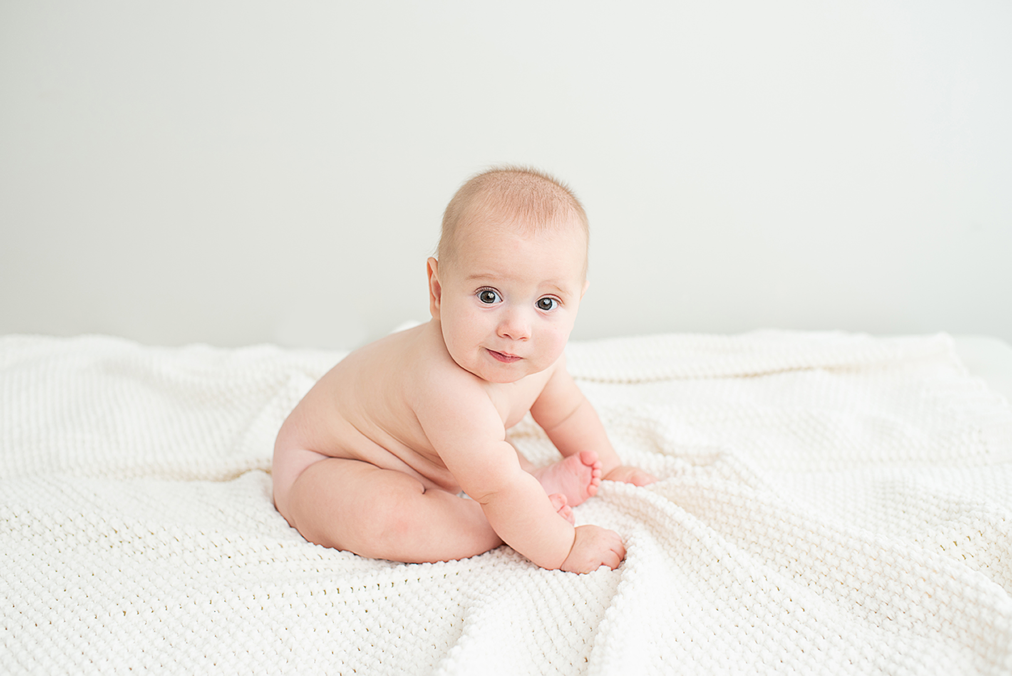 Smiling 6 month old baby sitting on a white blanket.