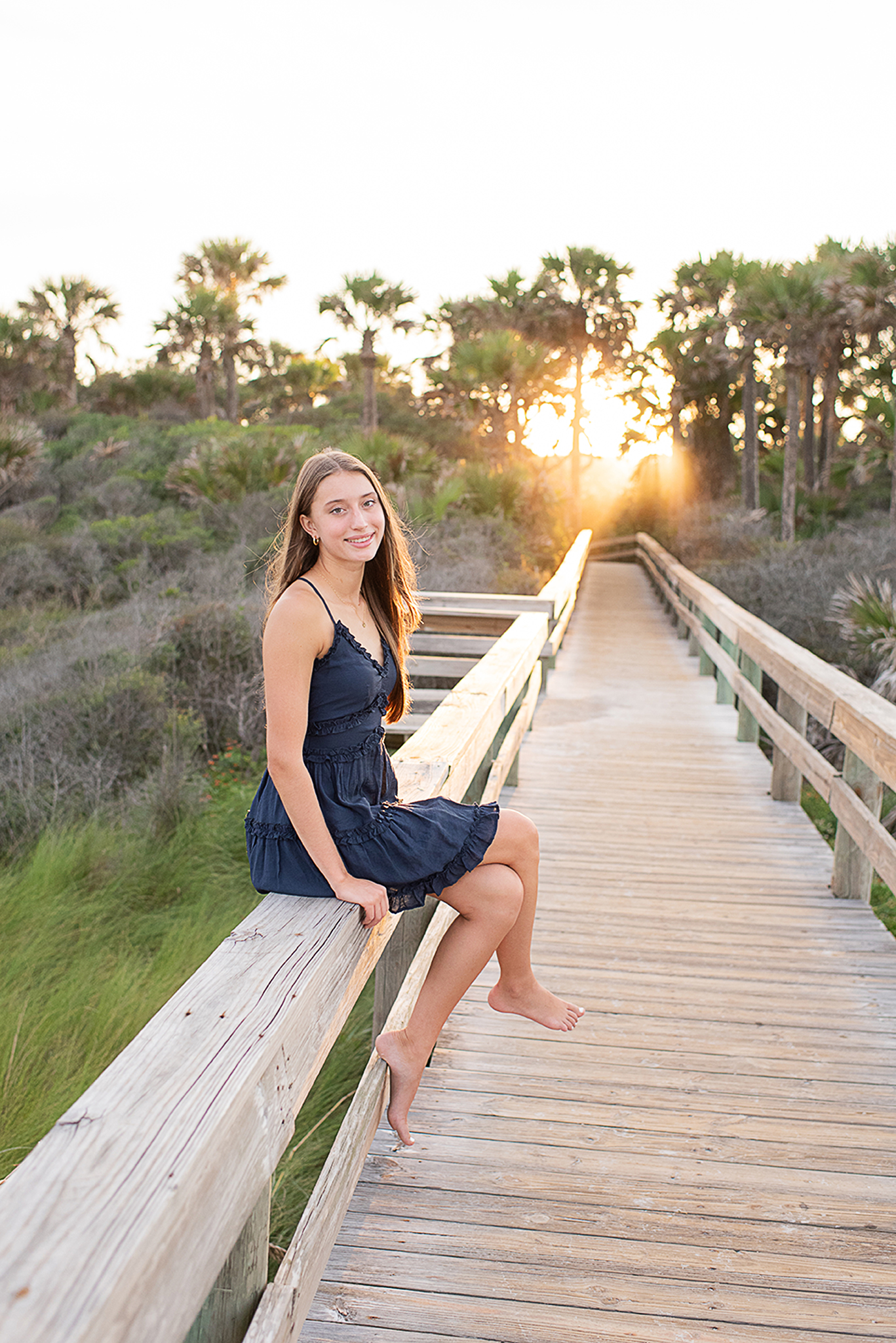 teenage girl sitting on pier at the beach with trees and sun behind her