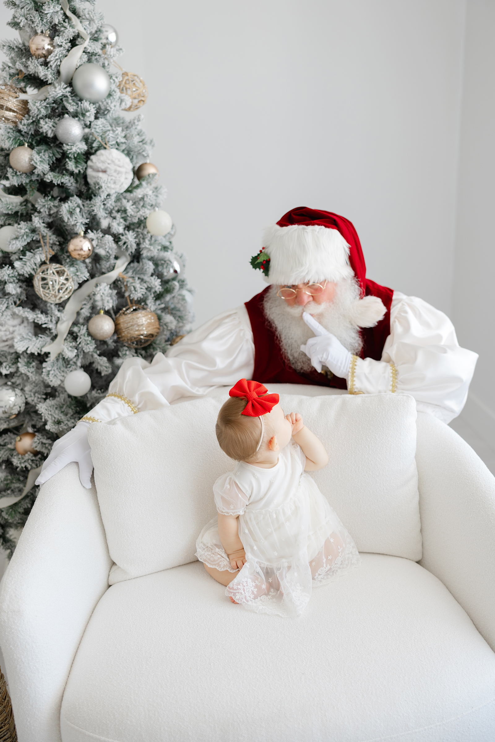 baby in white dress sitting on white chair with santa peeking behind her making the shhhh motion