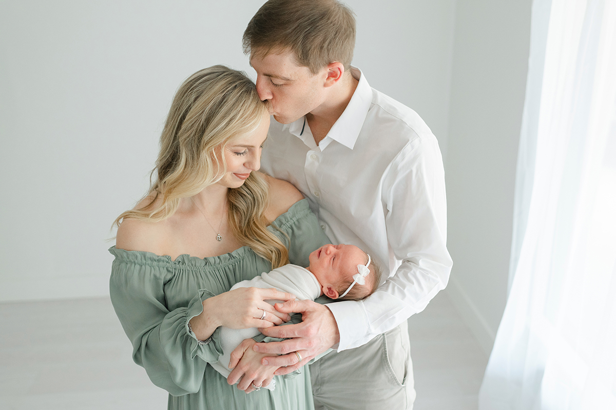 mom in green dress and dad in white shirt and khaki pants snuggling together with their new baby girl
