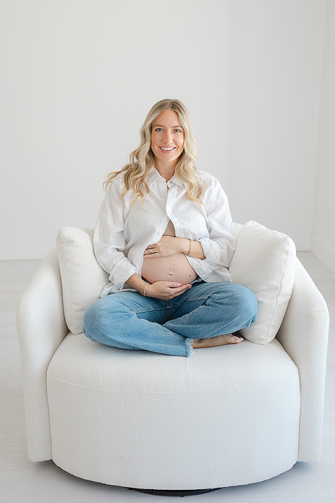 pregnant mom sitting on white chair with baby bump showing. shes in jeans and white button down shirt