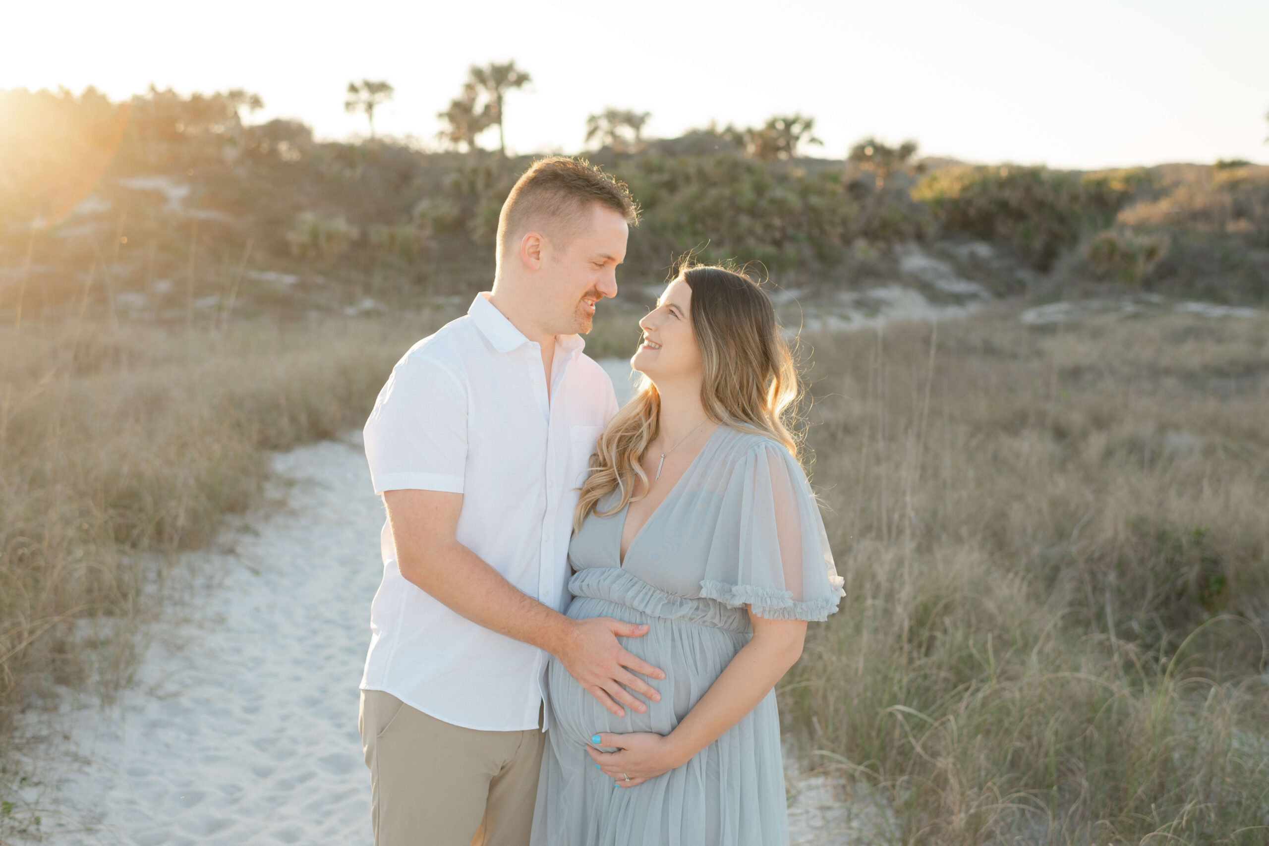 A happy pregnant would smile at each other while standing in white and blue on a beach at sunset