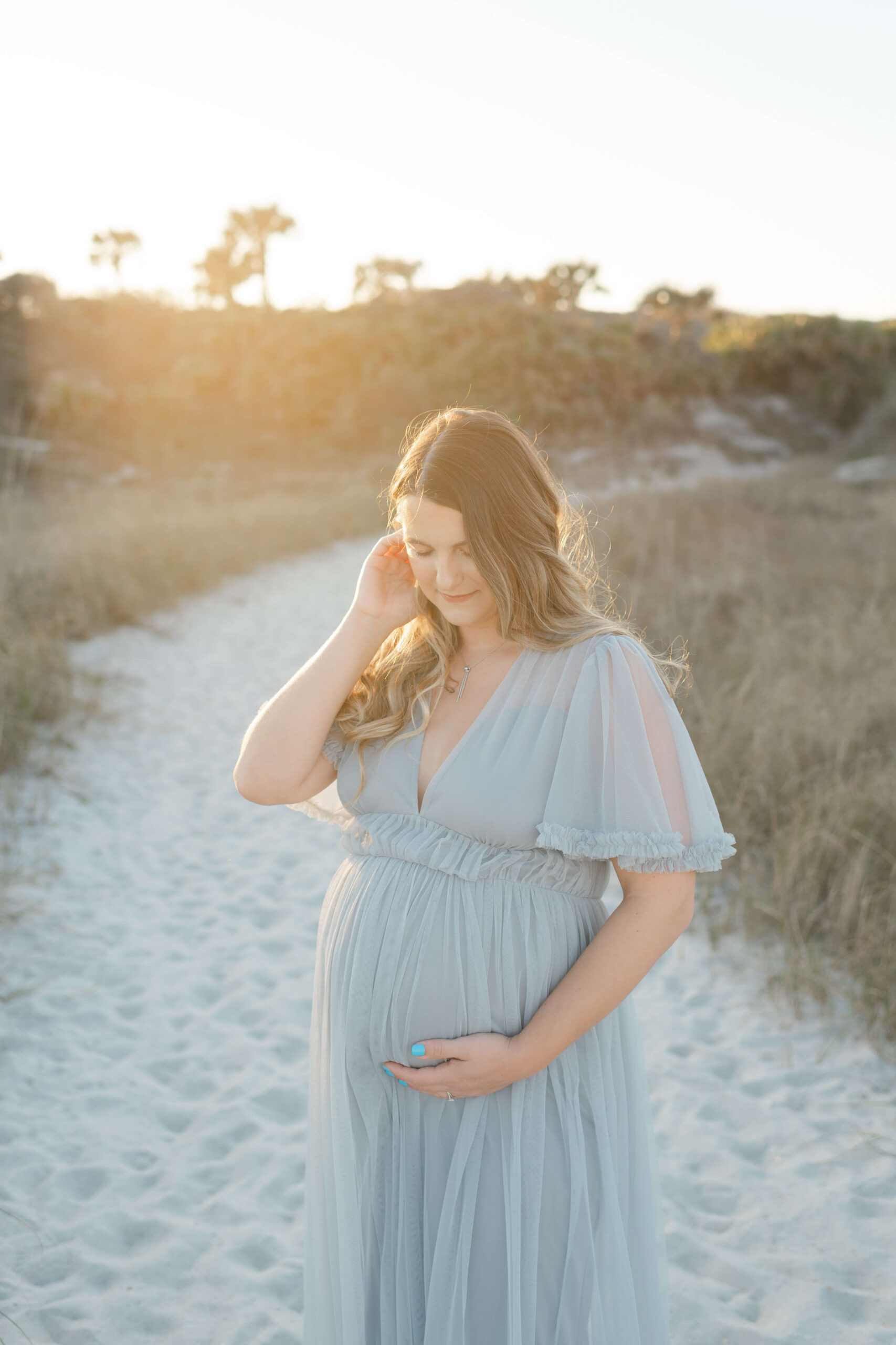 An expecting mother in a blue maternity gown pulls her hair back while smiling down to her bump on a beach trail at sunset thanks to IVF in Jacksonville, FL