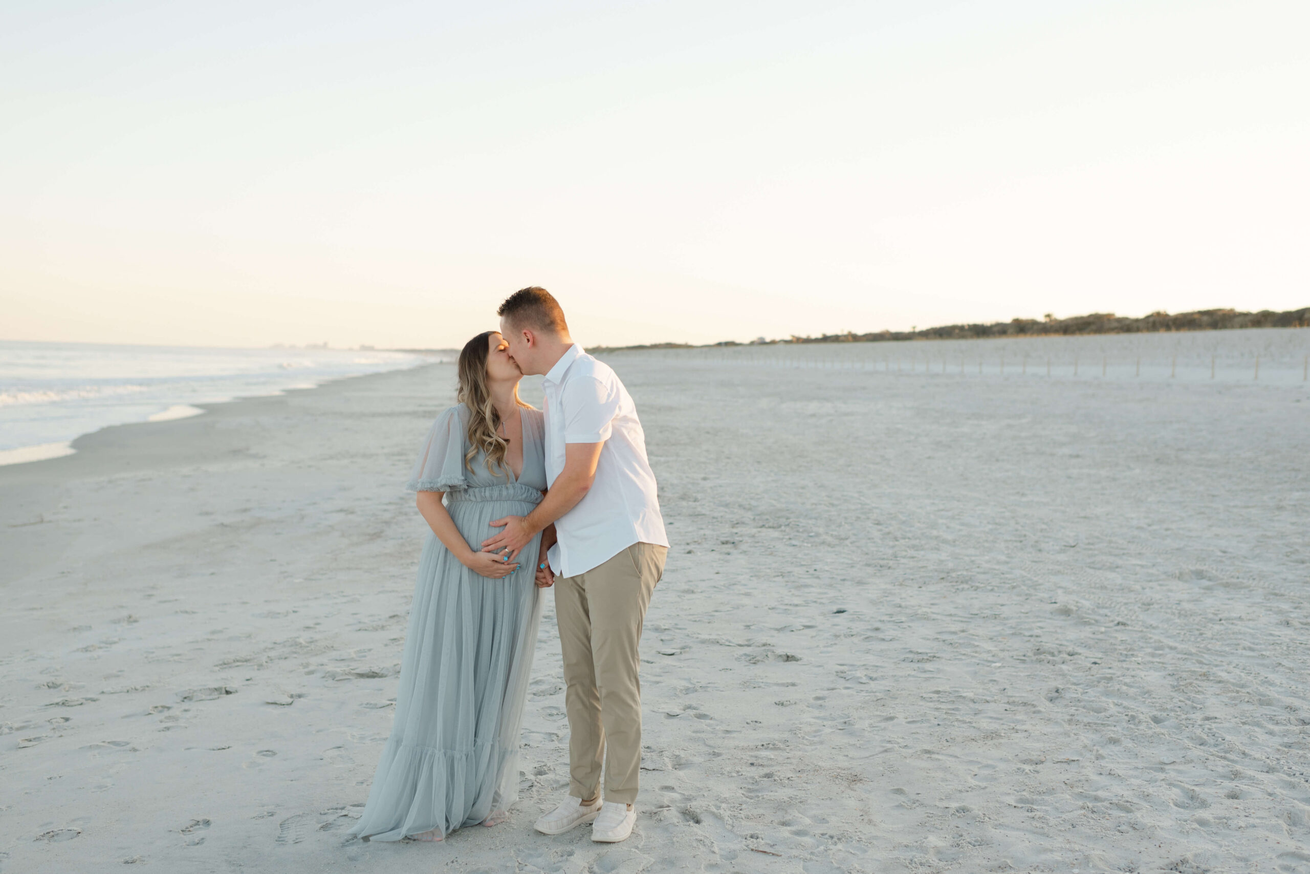 An expecting couple kisses on the beach at sunset in a white shirt and a blue maternity gown
