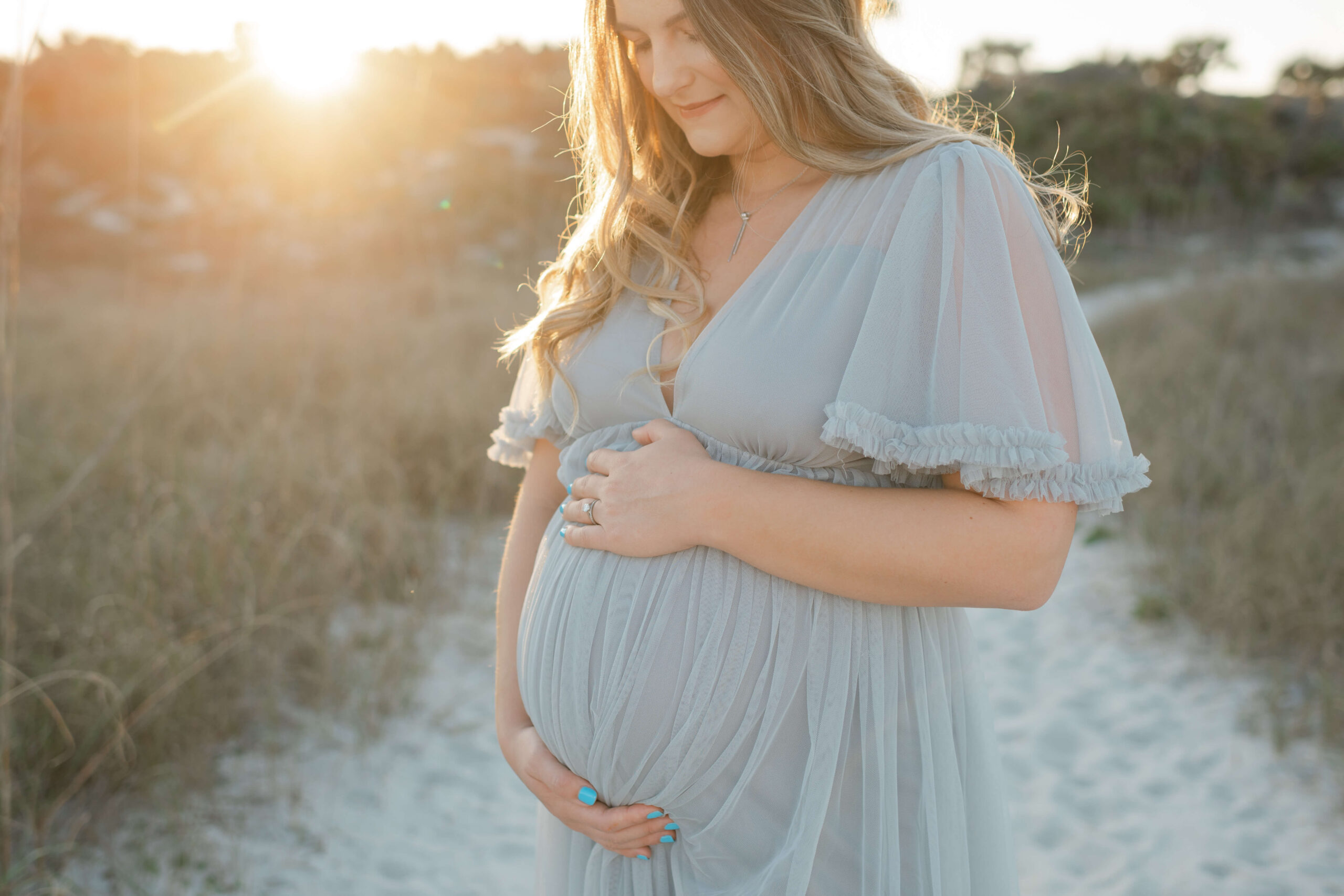 A pregnant woman smiles down to her hands on her bump in a blue maternity gown at sunset on a beach dune trail thanks to IVF in Jacksonville, FL