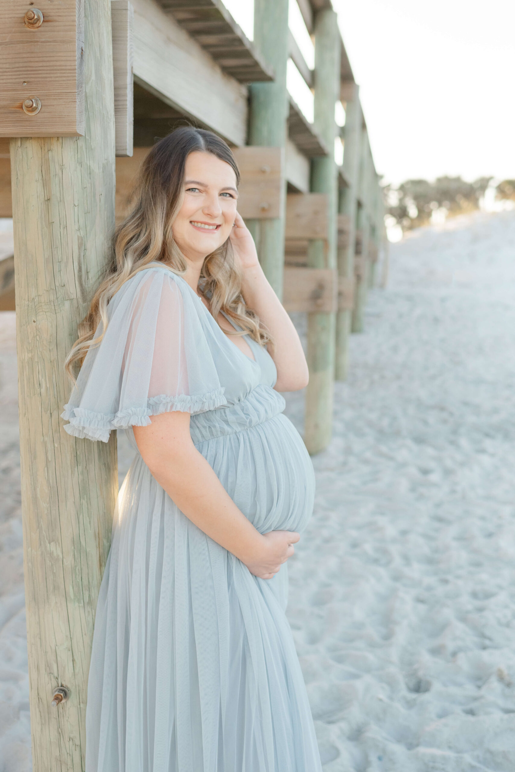 A happy mother to be in a blue maternity gown leans against a beach pier at sunset after finding success with IVF in Jacksonville, FL