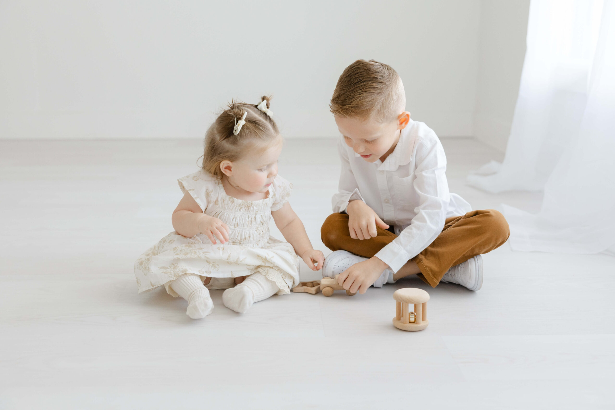 A toddler boy in a white shirt and brown pants plays with wooden toys with his younger sister on the studio floor under a window