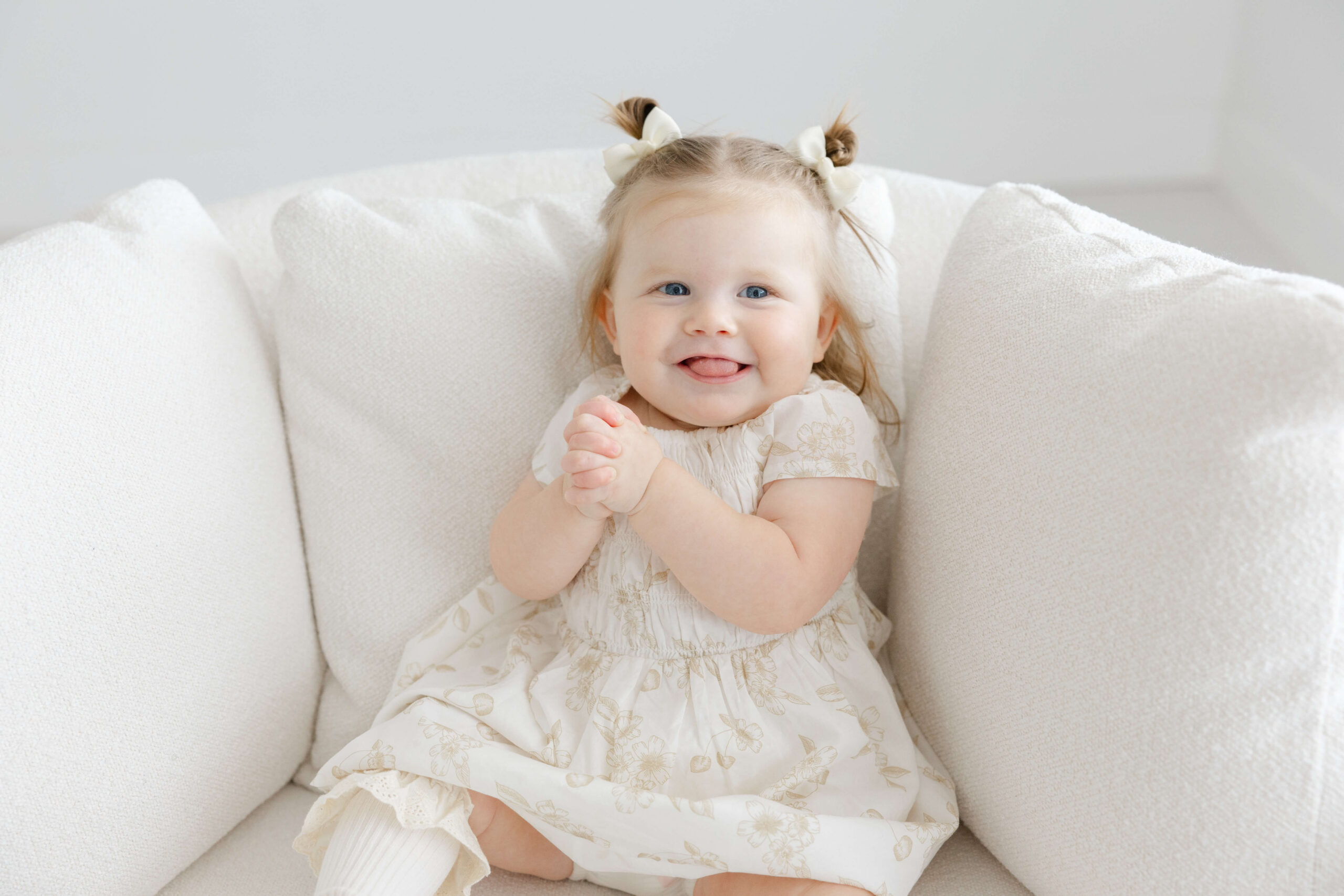 A happy toddler in a white dress sits in a couch in a studio clapping with a smile after visiting children's museums in Jacksonville, FL
