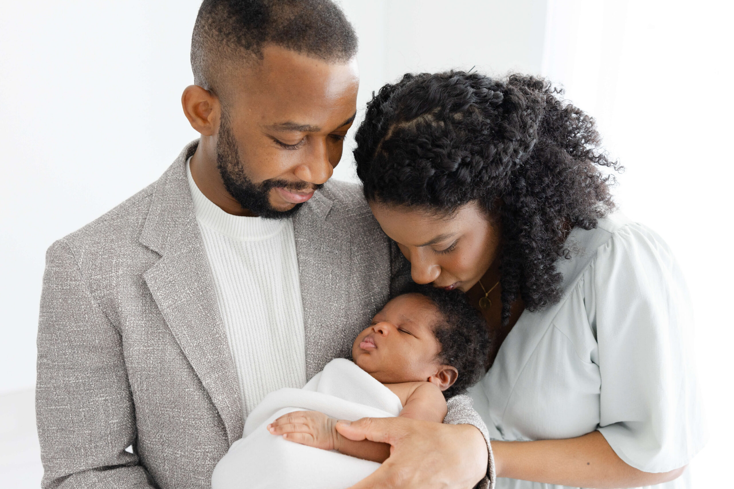 A mom leans down to kiss the head of her sleeping newborn in dad's arms