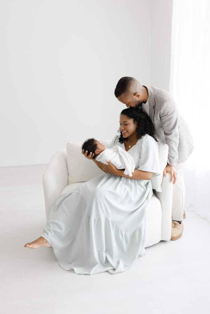 Infant photography outfit ideas with a mother in a light blue dress and father in neutral clothing posing with their newborn in a bright studio.