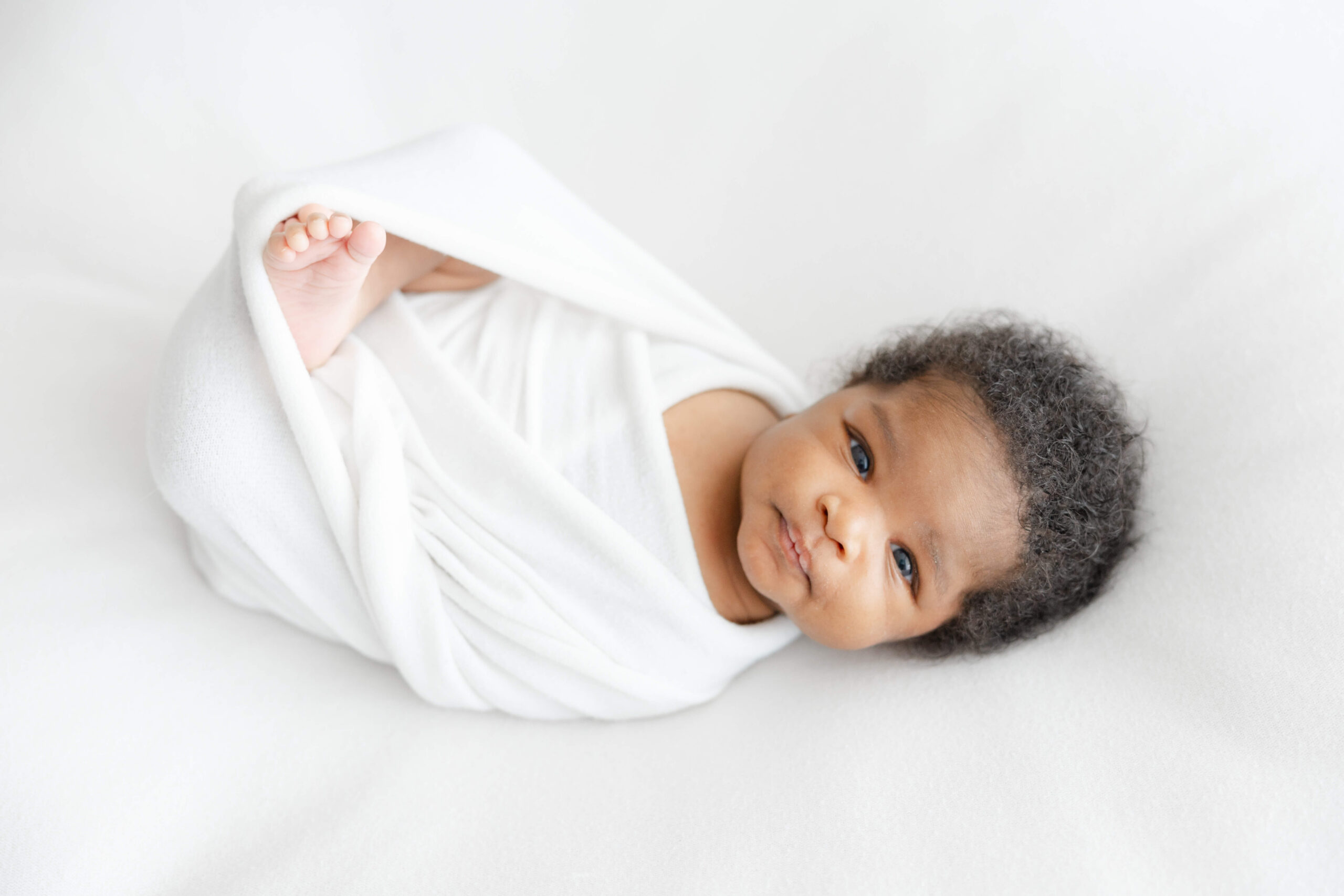 A newborn baby in a white swaddle lays with eyes open on a bed after some infant swim lessons in Jacksonville, FL