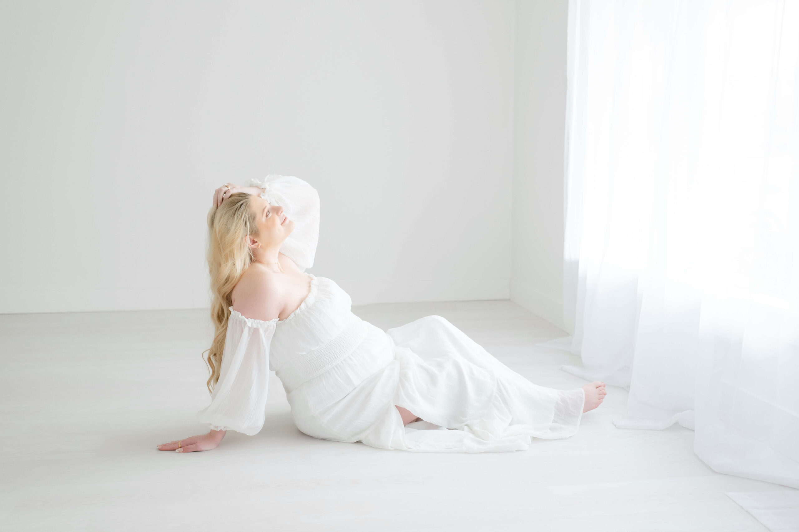 A smiling pregnant woman relaxes across the floor of a studio under a window with a hand in her hair in a white maternity dress