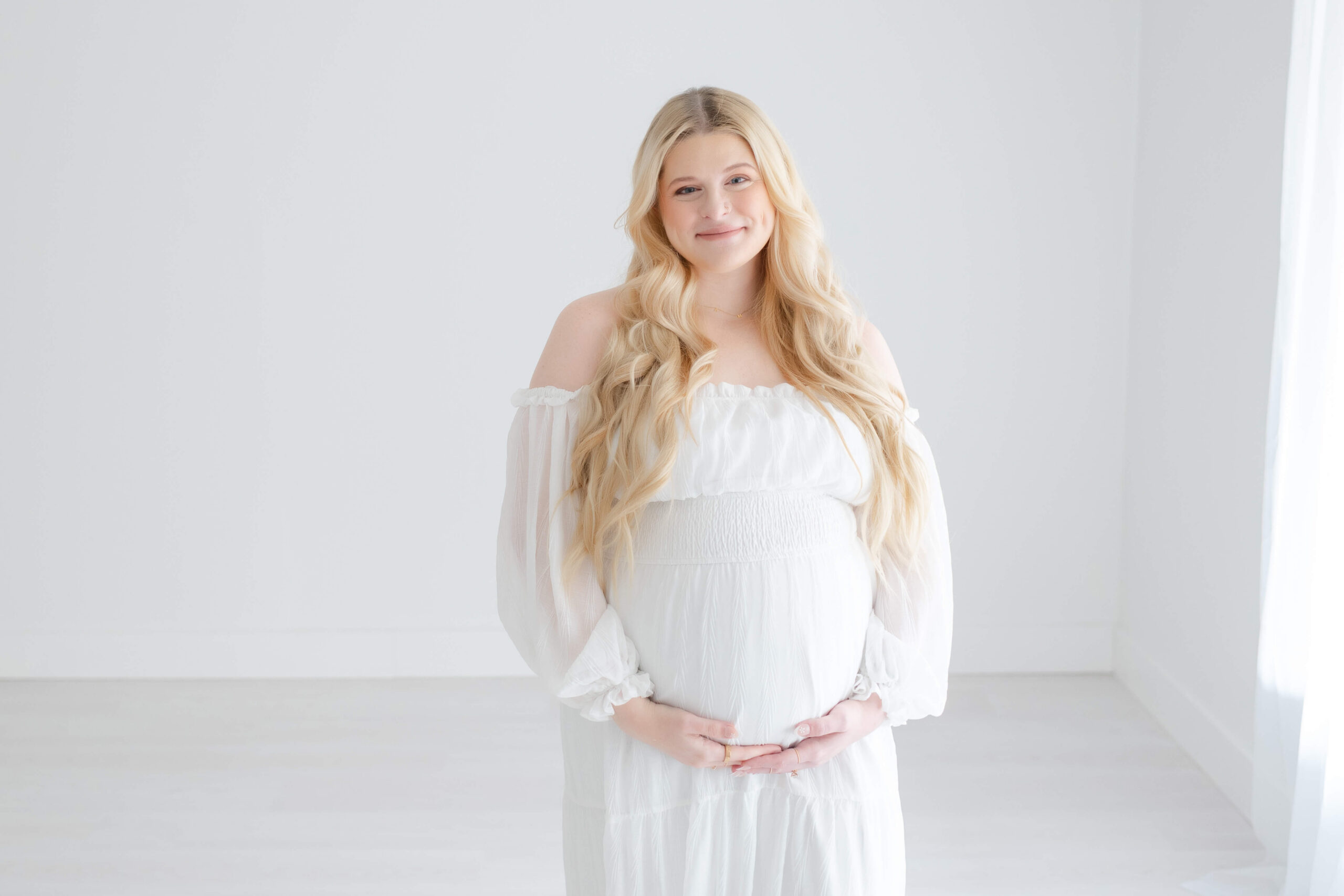 A blonde and smiling woman during pregnancy stands in a studio in a window with hands under her bump after meeting obgyns in Jacksonville, FL