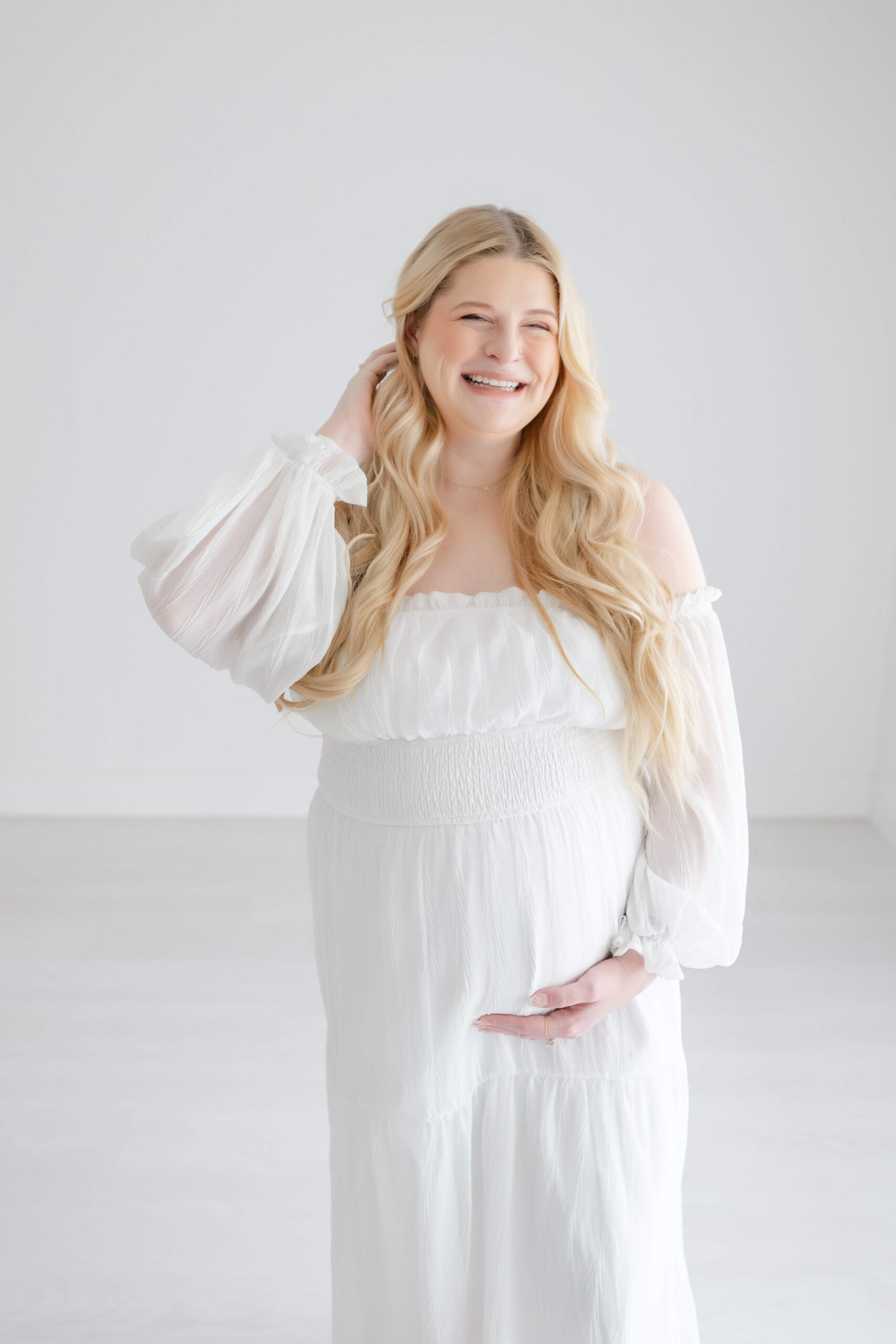 A giggling mom to be holds her bump while wearing a white maternity gown in a studio and pulling her hair behind her ear