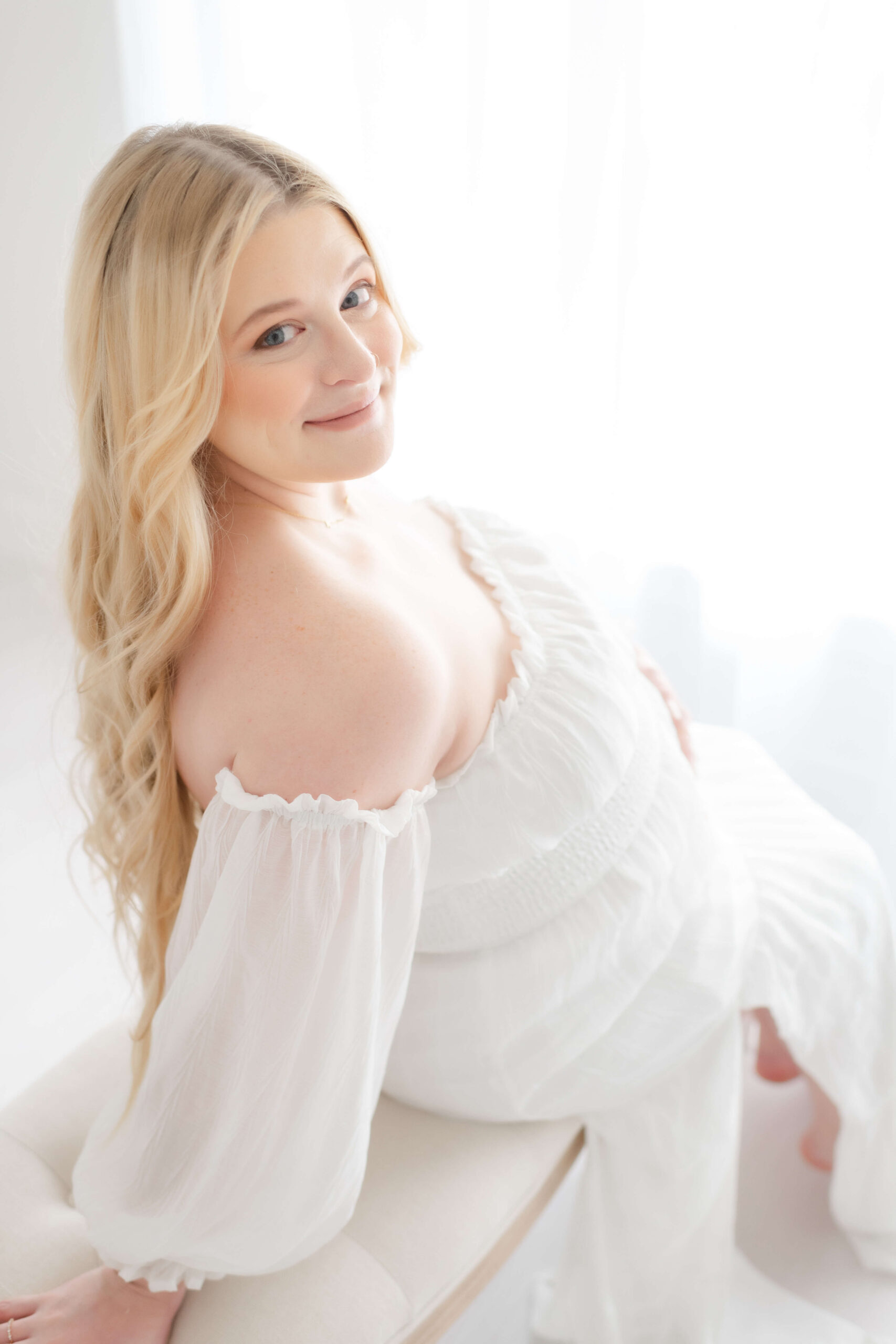 A smiling mother to be in a white off the shoulder maternity dress sits on a bench smiling back over her shoulder after finding obgyns in Jacksonville, FL