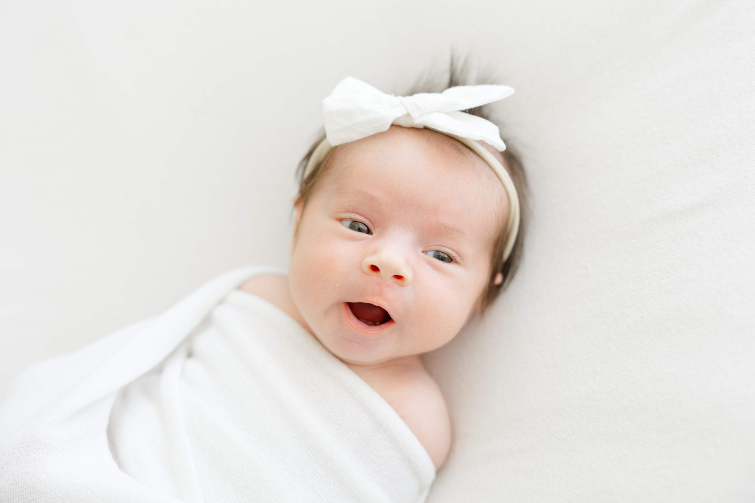 A newborn baby yawns while laying on a white bed in a matching swaddle